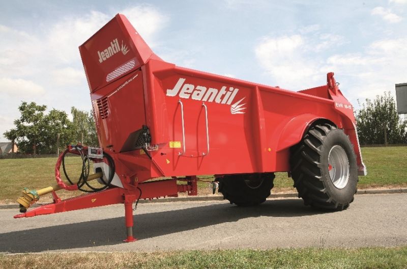 Une remorque agricole rouge, étiquetée « Jeantil », est garée sur une route pavée. Cet épandeur à fumier Jeantil est doté de gros pneus noirs et est encadré par un arrière-plan d'herbe et d'arbres.