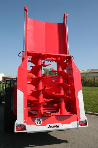 Une grande machine agricole rouge, ressemblant à un épandeur à fumier Jeantil, est garée sur une route. Elle est équipée de vis sans fin et affiche le numéro 25 sur un panneau à l'arrière, située près d'une zone herbeuse sous un ciel bleu clair.