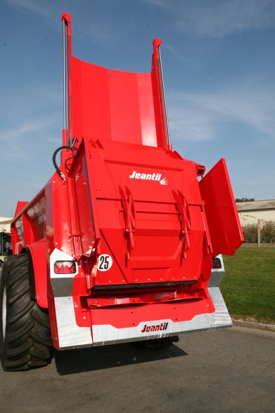 Un épandeur à fumier Jeantil rouge vif avec de gros pneus est garé sur une route pavée. Le nom de la marque « Jeantil » est visible à l'arrière. Le ciel est clair et bleu, avec de l'herbe verte bordant le bord de la route.