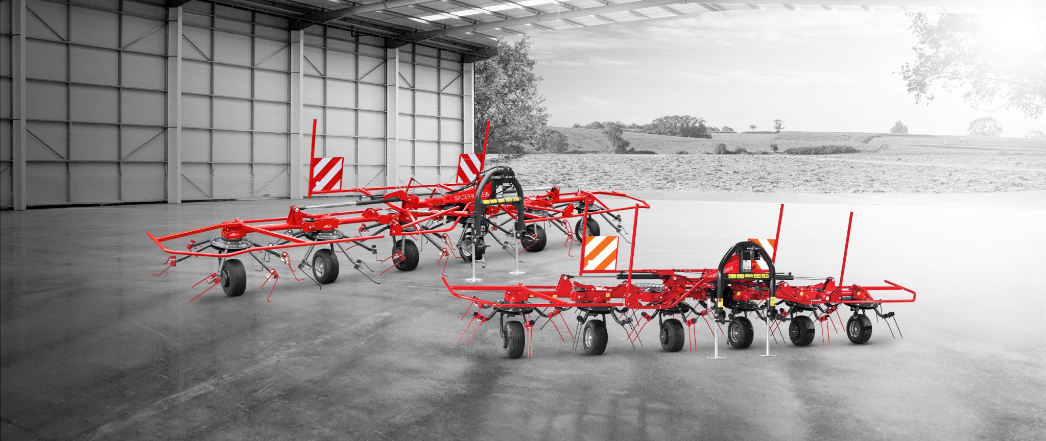 Trois machines agricoles rouges sont garées dans un grand entrepôt ouvert, avec vue sur un paysage rural en arrière-plan. Les machines sont dotées de plusieurs roues et de composants rotatifs.