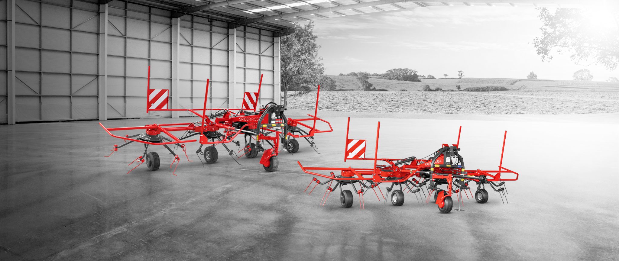 Des machines agricoles rouges sont exposées dans un grand garage ouvert. Trois machines sont disposées sur un sol en béton poli avec un vaste paysage rural visible en arrière-plan à travers la porte ouverte.