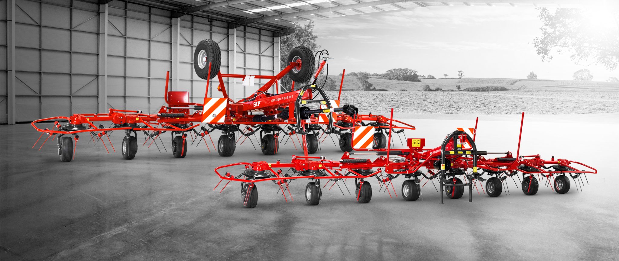 Une collection de machines agricoles rouges, dont une faneuse et un râteau, est exposée dans un grand entrepôt intérieur. L'espace ouvert révèle des champs et des arbres en arrière-plan à travers le côté ouvert du bâtiment.