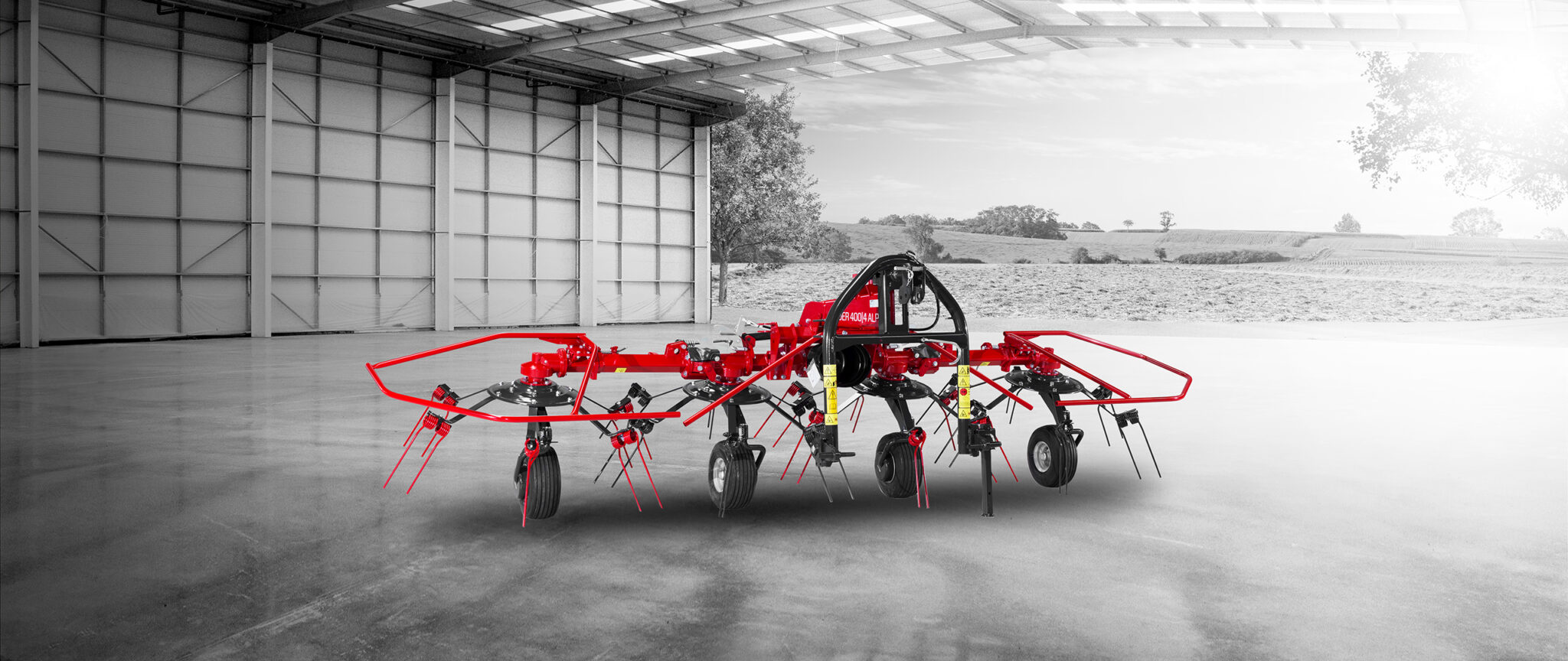 Une machine agricole rouge est positionnée à l'intérieur d'un entrepôt avec une vue dégagée sur les champs extérieurs. La machine est dotée de plusieurs dents et roues, et l'arrière-plan est principalement en niveaux de gris, mettant en valeur la couleur rouge vibrante.