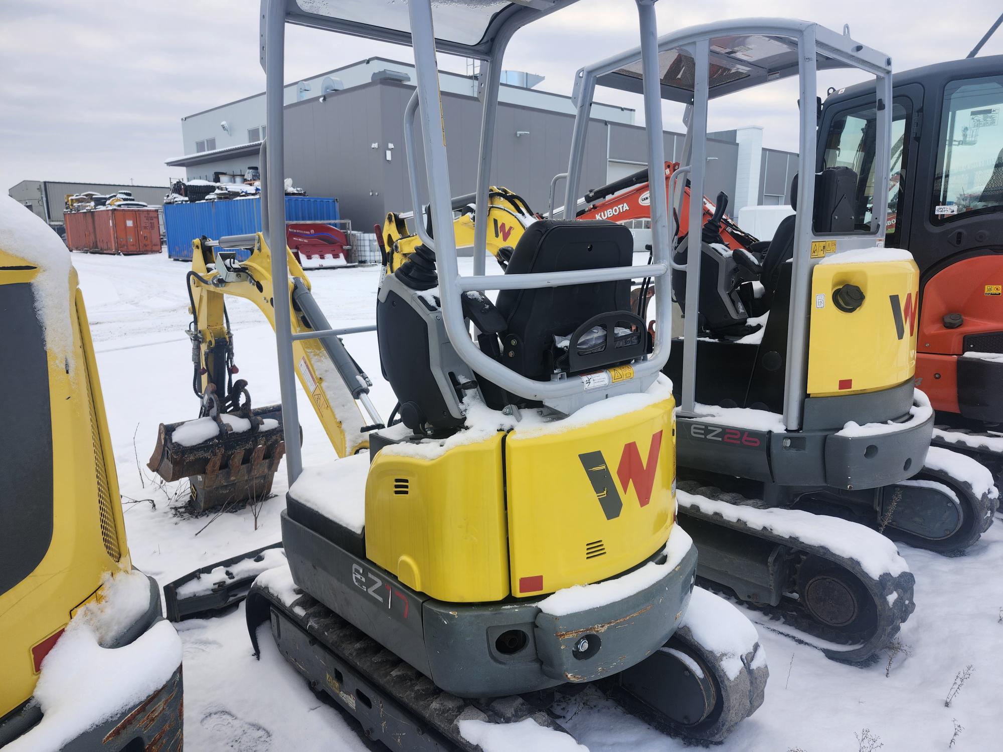 Des excavatrices compactes jaunes recouvertes de neige sont alignées sur un site industriel. Un bâtiment gris et des conteneurs de stockage sont visibles en arrière-plan.