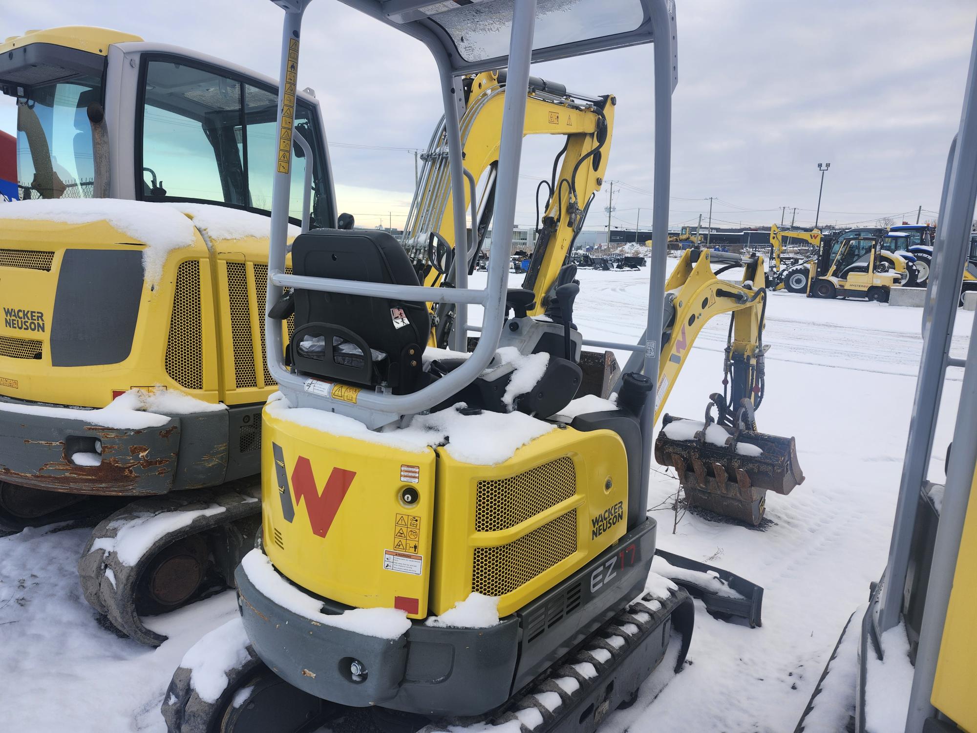 Une mini-pelle jaune recouverte de neige est garée à côté d'un autre véhicule de chantier sur un terrain enneigé. Le godet de la pelle est orienté vers le bas et le ciel en arrière-plan est nuageux.