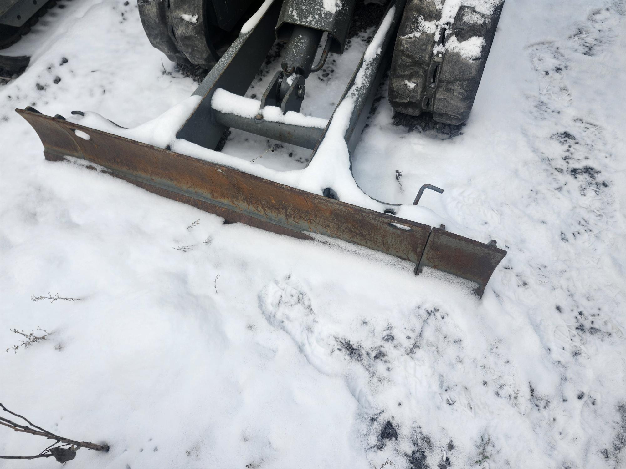 Un chasse-neige pour véhicule est posé au sol, recouvert de neige. La lame en métal rouillée est partiellement enfouie dans la neige, avec des traces de pas laissées par un véhicule relié à elle visibles derrière elle. Des branches nues clairsemées sont visibles au premier plan.