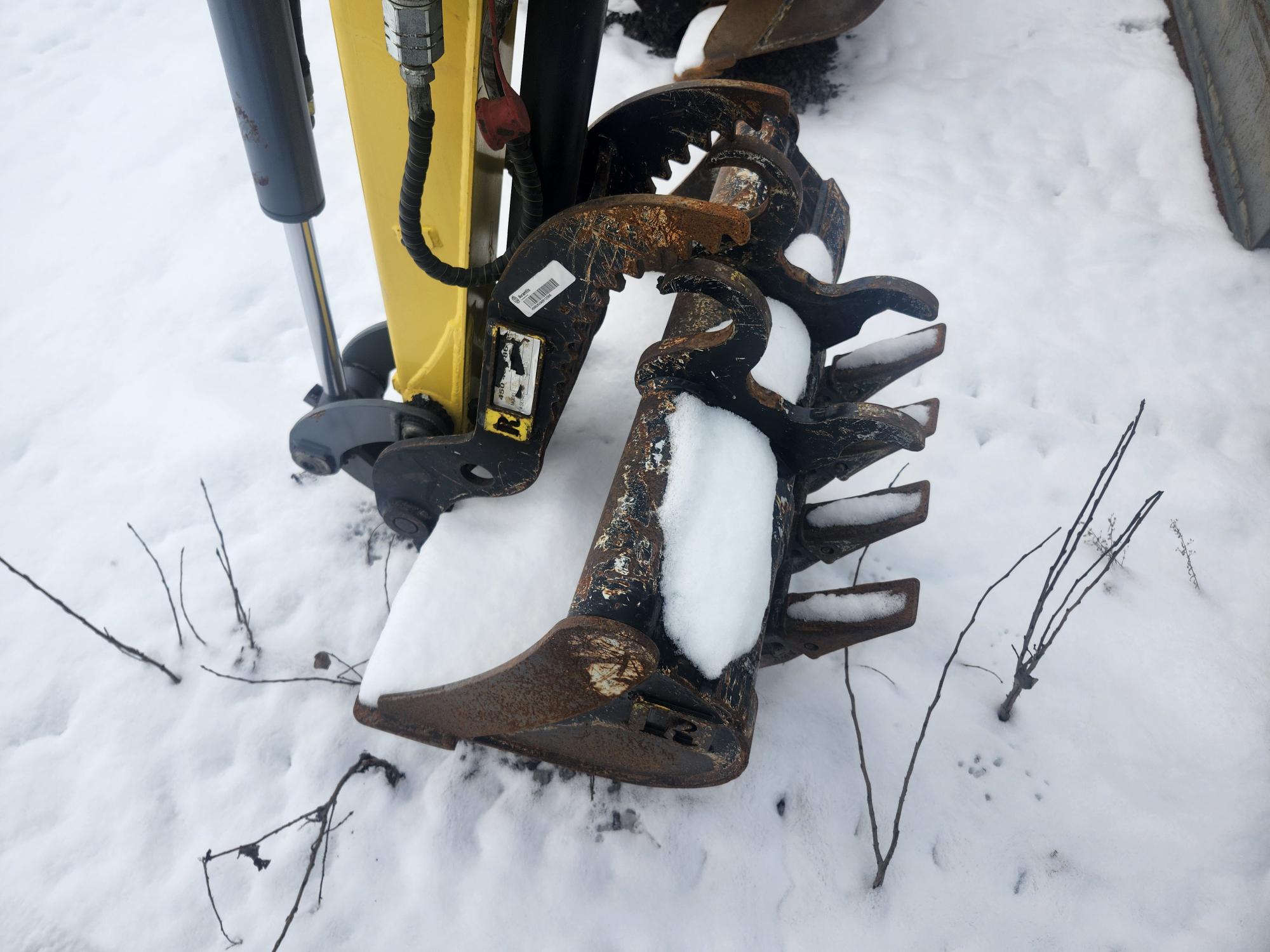 Une griffe d'excavatrice rouillée, partiellement recouverte de neige, repose sur un sol enneigé. De fines brindilles dénudées sont visibles à proximité. Le bras hydraulique est à l'arrière-plan, contrastant avec l'environnement blanc.