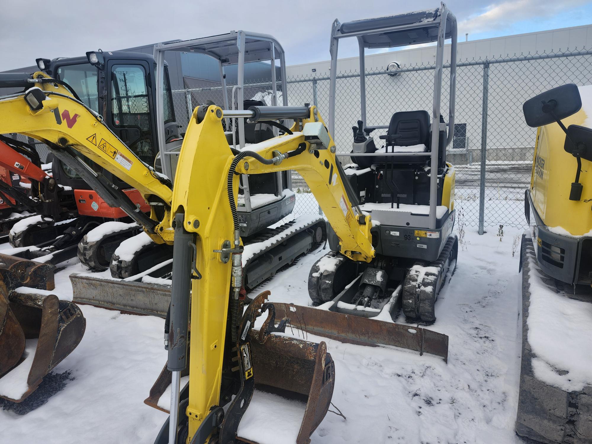 Une rangée de mini-pelles jaunes recouvertes d'une fine couche de neige est garée à l'extérieur. Elles sont alignées les unes à côté des autres sur un sol enneigé, entouré d'une clôture grillagée. Un bâtiment est visible en arrière-plan sous un ciel nuageux.