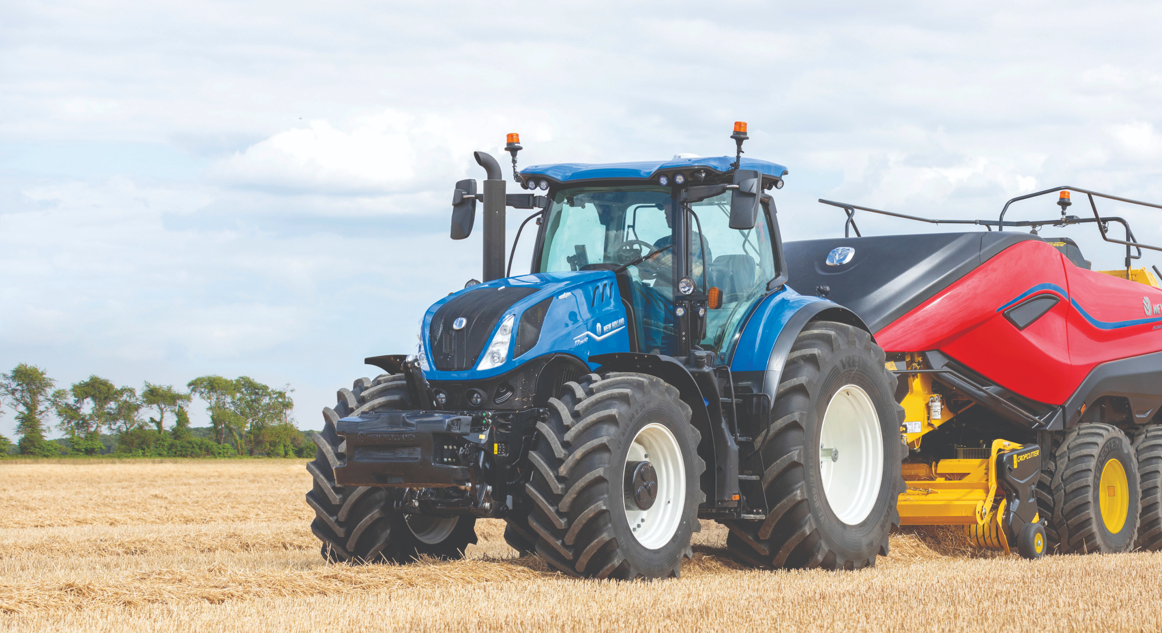 Un tracteur bleu travaille dans un champ, attaché à une grosse machine agricole rouge et jaune. Les gros pneus et la construction puissante du tracteur suggèrent qu'il est utilisé pour des tâches agricoles lourdes. L’arrière-plan montre un vaste champ avec quelques arbres au loin.