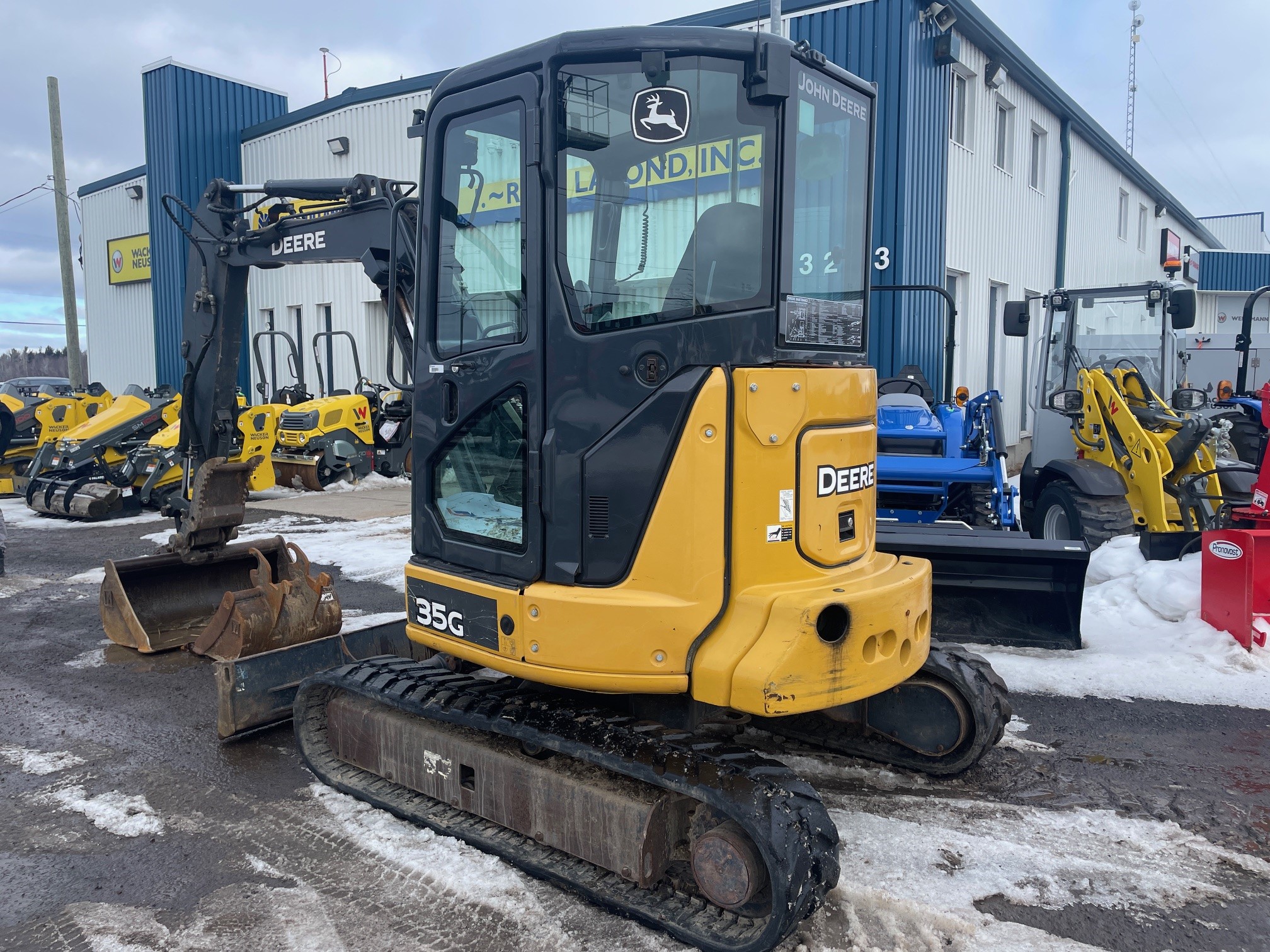 A yellow and black John Deere 35G excavator parked in a snowy outdoor lot, in front of a blue and white building with the sign 