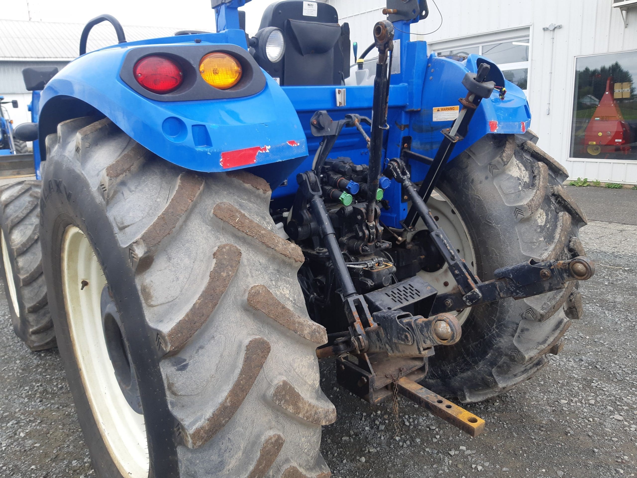 Vue arrière d'un tracteur bleu sale garé sur du gravier, montrant de gros pneus arrière usés, un attelage trois points et divers composants hydrauliques. Un bâtiment avec une fenêtre est visible en arrière-plan, reflétant les arbres et le ciel.