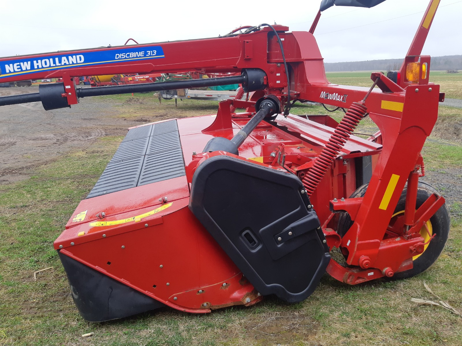 A New Holland Discbine 313, a red agricultural machine, is positioned on a grassy field. The machine features large cutting discs and a lifting mechanism. The brand and model name are visible on the equipment.