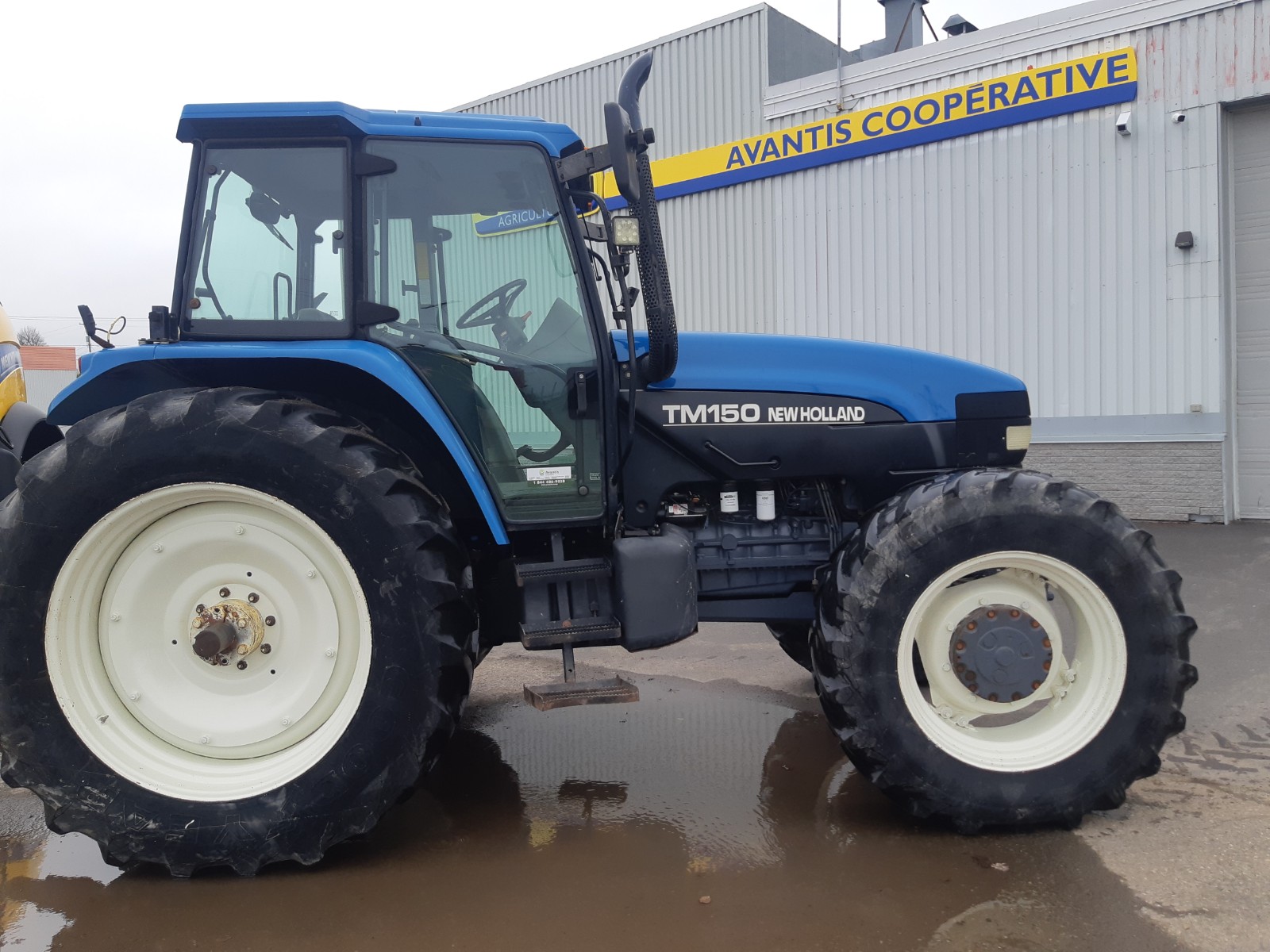 A blue New Holland TM150 tractor with large black tires is parked on wet ground in front of a gray building. The building has 