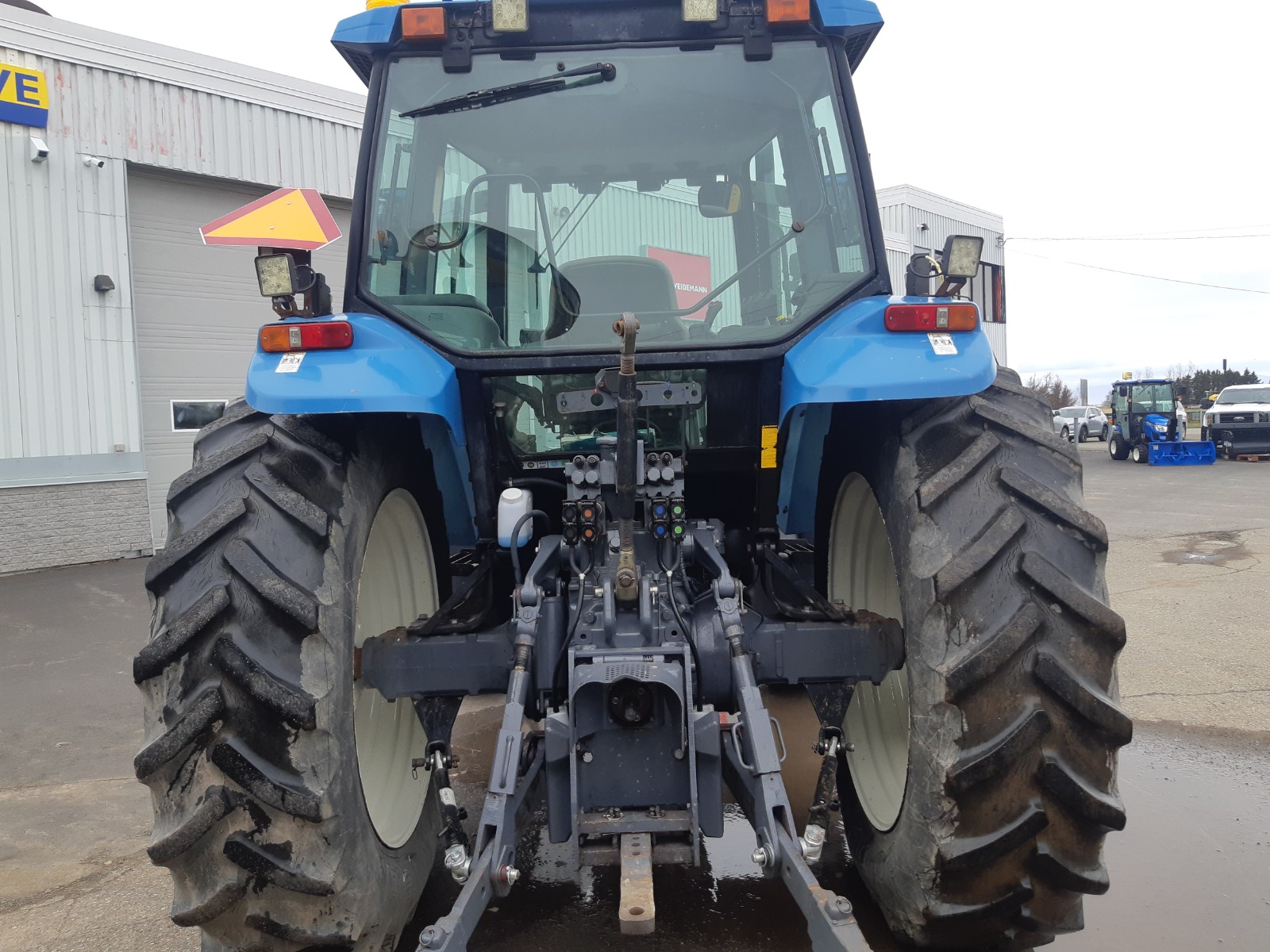 The image shows the rear view of a blue agricultural tractor with large tires and various hydraulic connections and linkages. It is parked on a concrete surface near a commercial building with a partially visible sign.