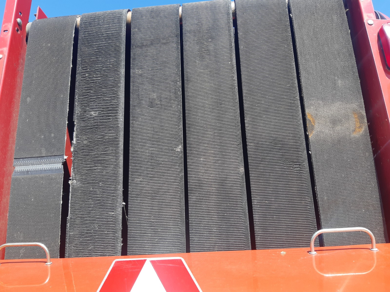 Close-up of a large industrial conveyor belt system with multiple black belts aligned vertically, mounted on a red framework. Parts of the red metal structure are visible on the sides, and a warning sign with a red and white reflective triangle can be seen at the bottom.