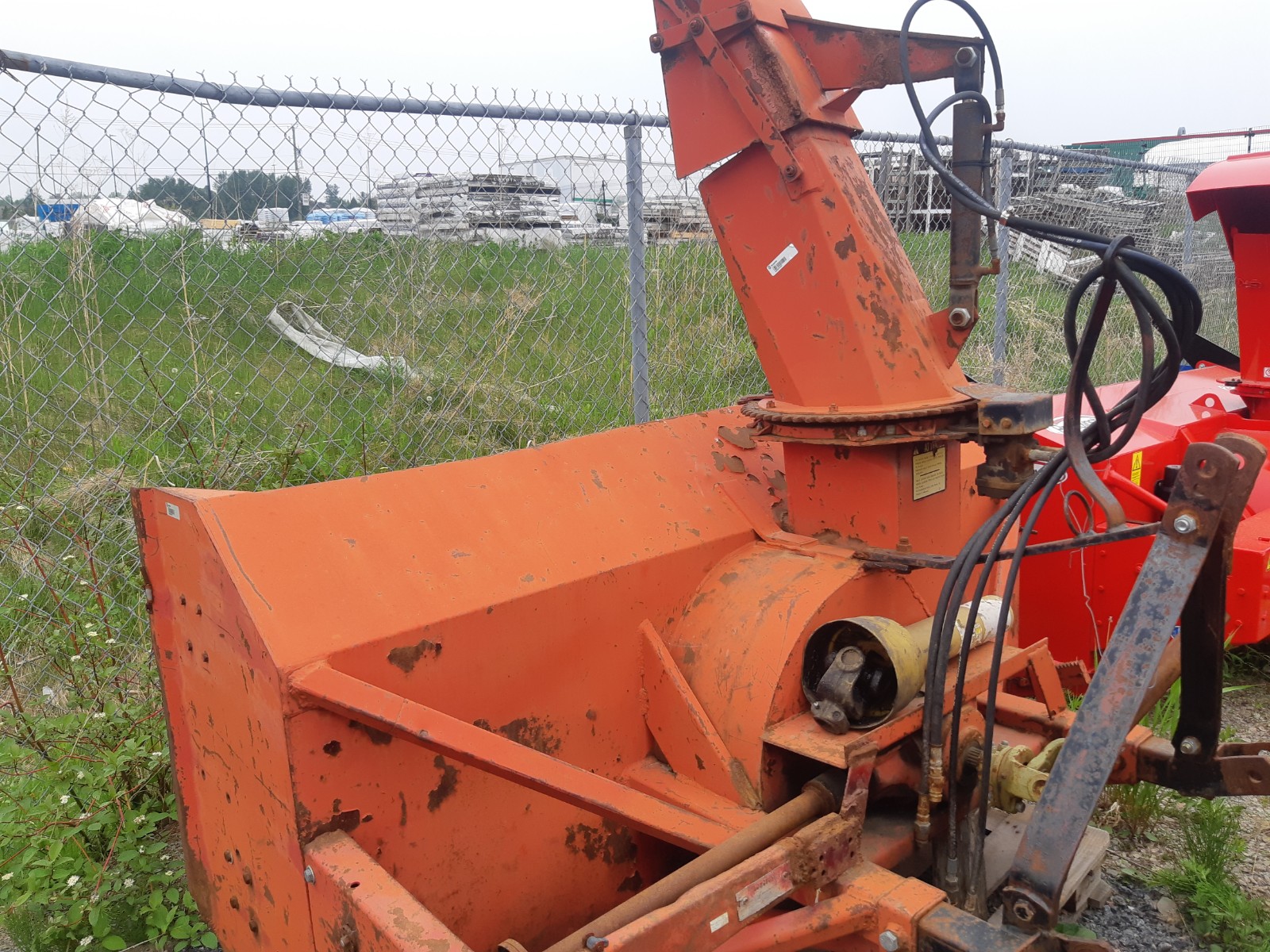 An orange snow blower attachment connected to a piece of machinery sits next to a chain-link fence in a grassy area. The snow blower is weathered, showing signs of rust and wear. An industrial area with various equipment is visible in the background.