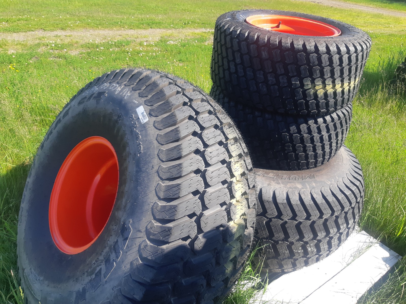 A stack of four large tires with bright red rims sits on grassy ground. Three of the tires are stacked vertically, while one is leaning against the stack. The background shows more grass and a dirt path.