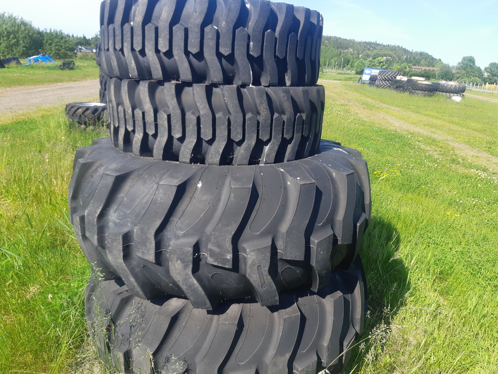 Stack of large, heavy-duty tires on green grass with additional tires and a partly cloudy blue sky visible in the background. The tires feature deep, rugged treads and are arranged in two stacks, each consisting of four tires.