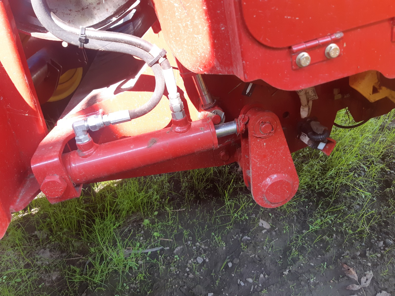 Close-up of the hydraulic components of a red farming machine. The image shows multiple hoses, bolts, and a hydraulic cylinder connected to the machine, which is positioned over grass and dirt. The bright red color of the machinery contrasts with the green grass below.