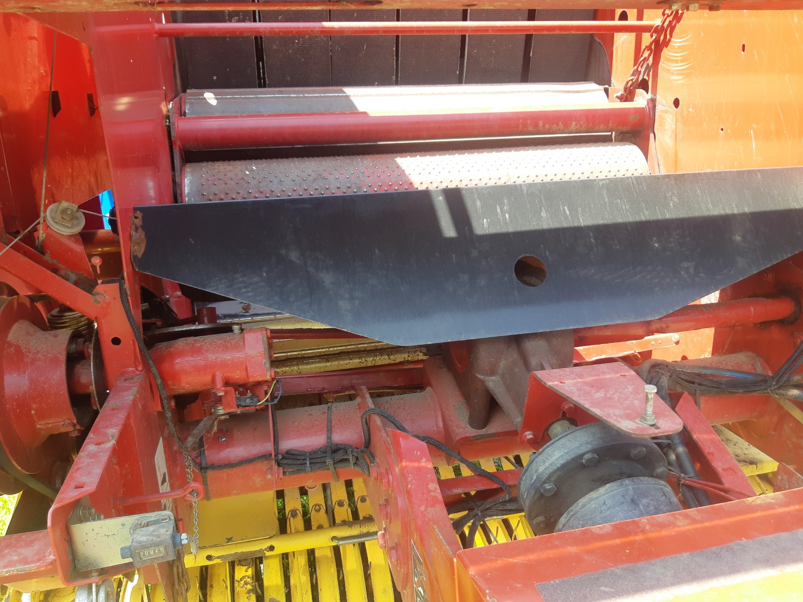 Close-up view of agricultural machinery, showing the internal components of a red combine harvester. Visible parts include metal rollers, gears, hoses, and a large black metal guard plate with a circular hole. The equipment appears well-used, with some wear marks.