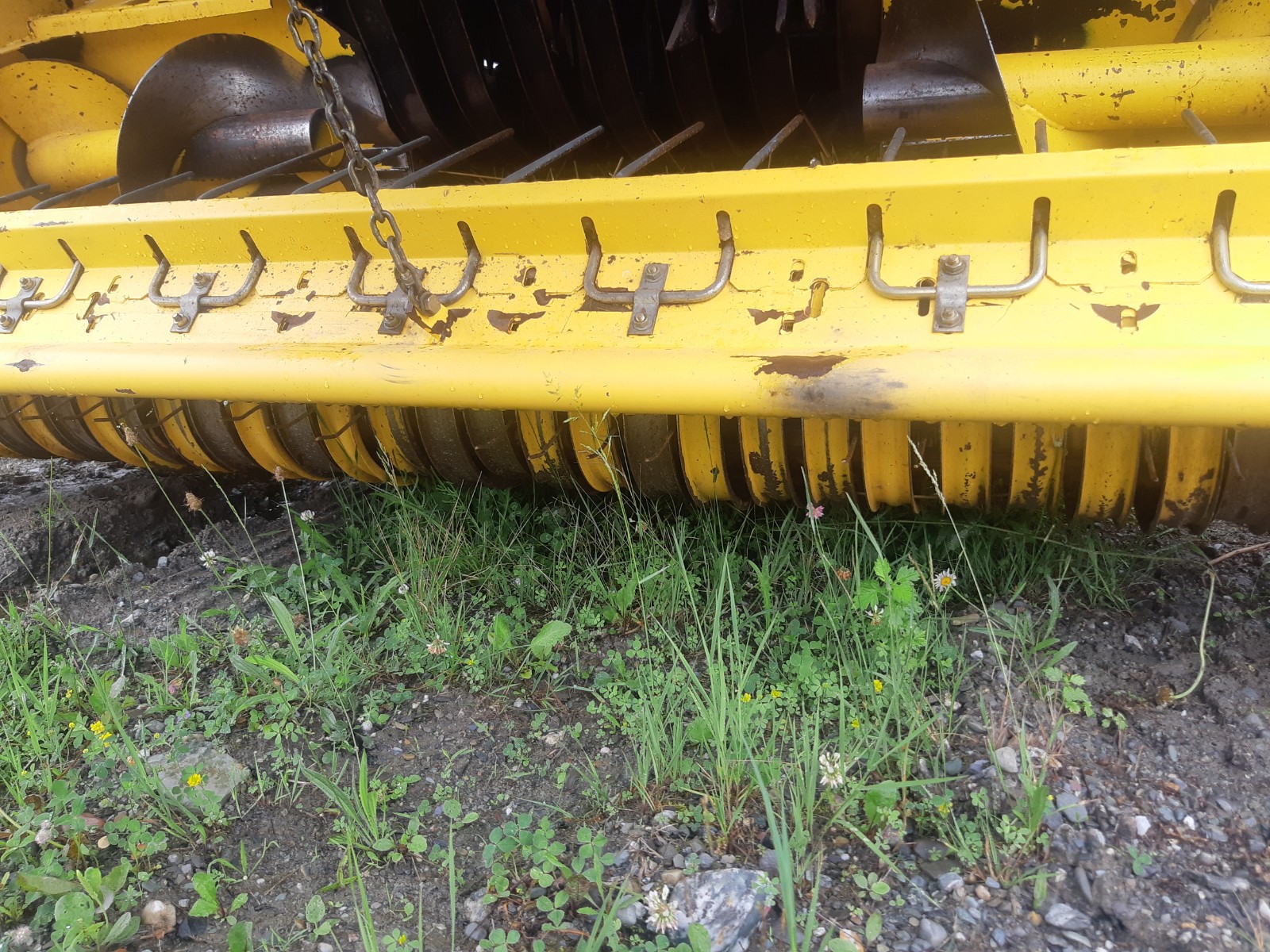 A close-up view of a yellow agricultural machine with metal prongs and a rotary component resting on grass and rocky soil. The machine shows signs of wear and use, with some rust and dirt visible on the equipment.