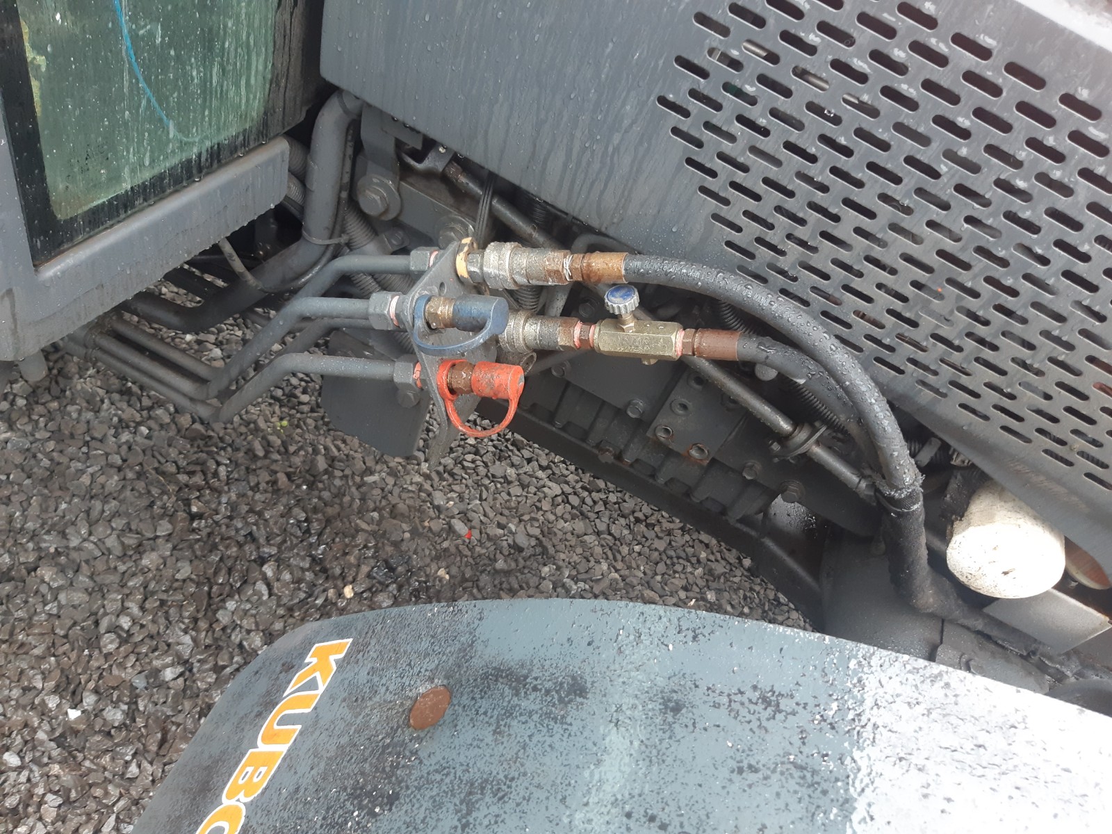 Close-up of hydraulic hoses and fittings on a piece of industrial machinery. The machinery is on a gravel surface, and part of the machinery has a metal grille. The Kubota logo can be seen on a metal component in the lower part of the image.