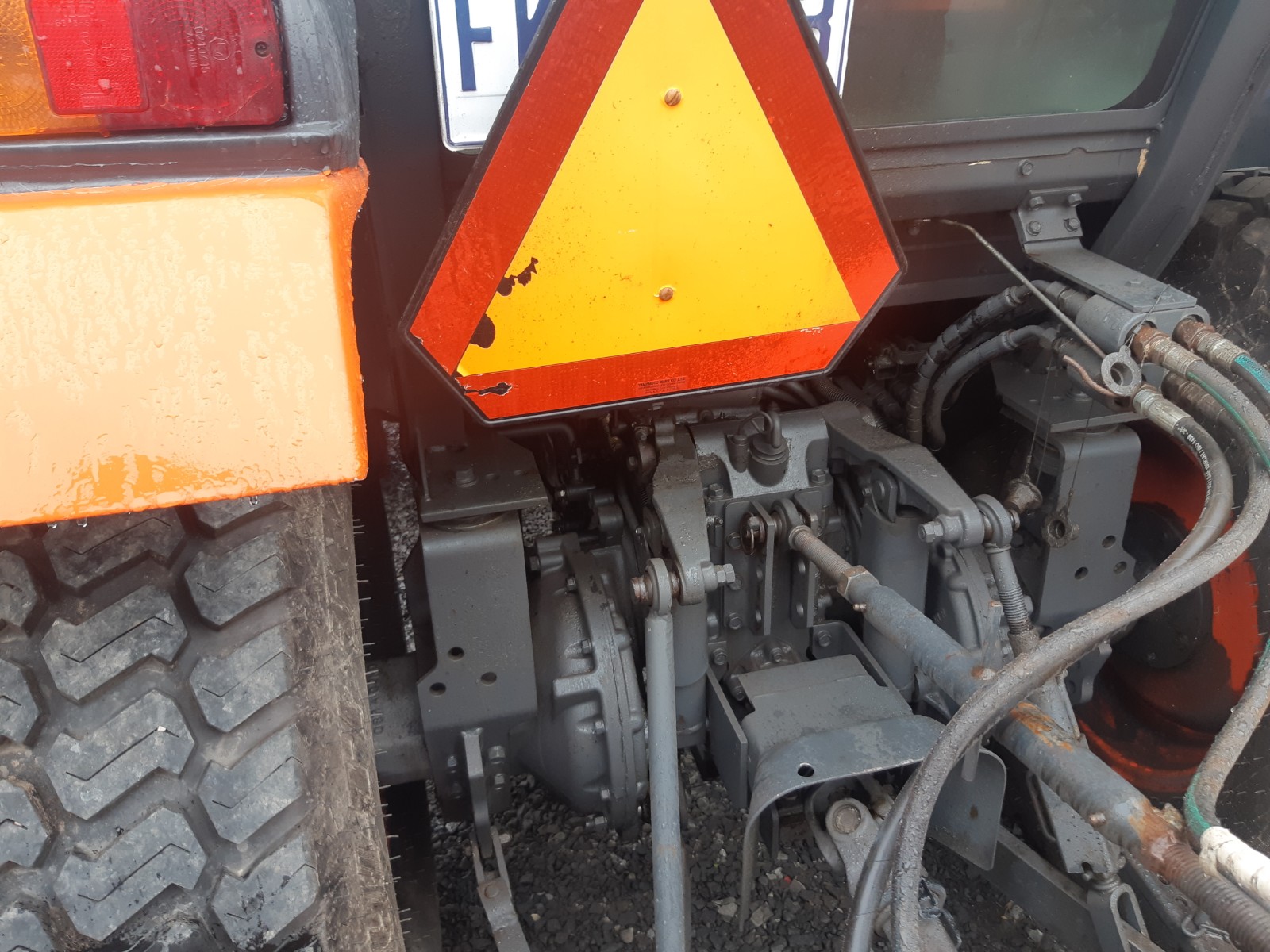 Close-up of the rear section of a tractor, featuring a bright orange hazard triangle sign mounted above mechanical parts and connections. The textured tire is visible on the left side. The equipment appears to be muddy, indicating recent use in wet conditions.