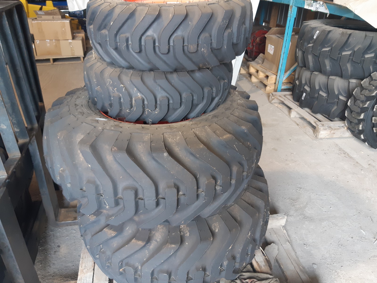 Stack of large, thick industrial tires with deep tread patterns are piled on wooden pallets in a warehouse. Shelves with various boxes and equipment can be seen in the background.