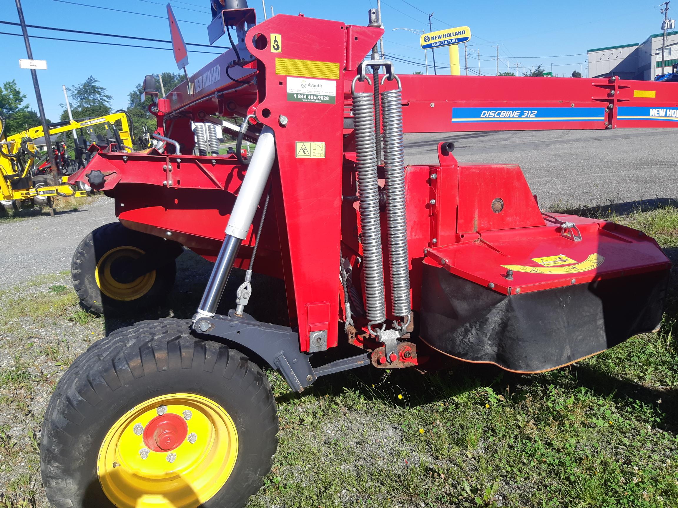 A bright red New Holland Discbine 312 agricultural machine is parked on gravel. It features large yellow wheels, visible spring mechanisms, and a mower deck. In the background are other agricultural equipment and a building under a clear blue sky.