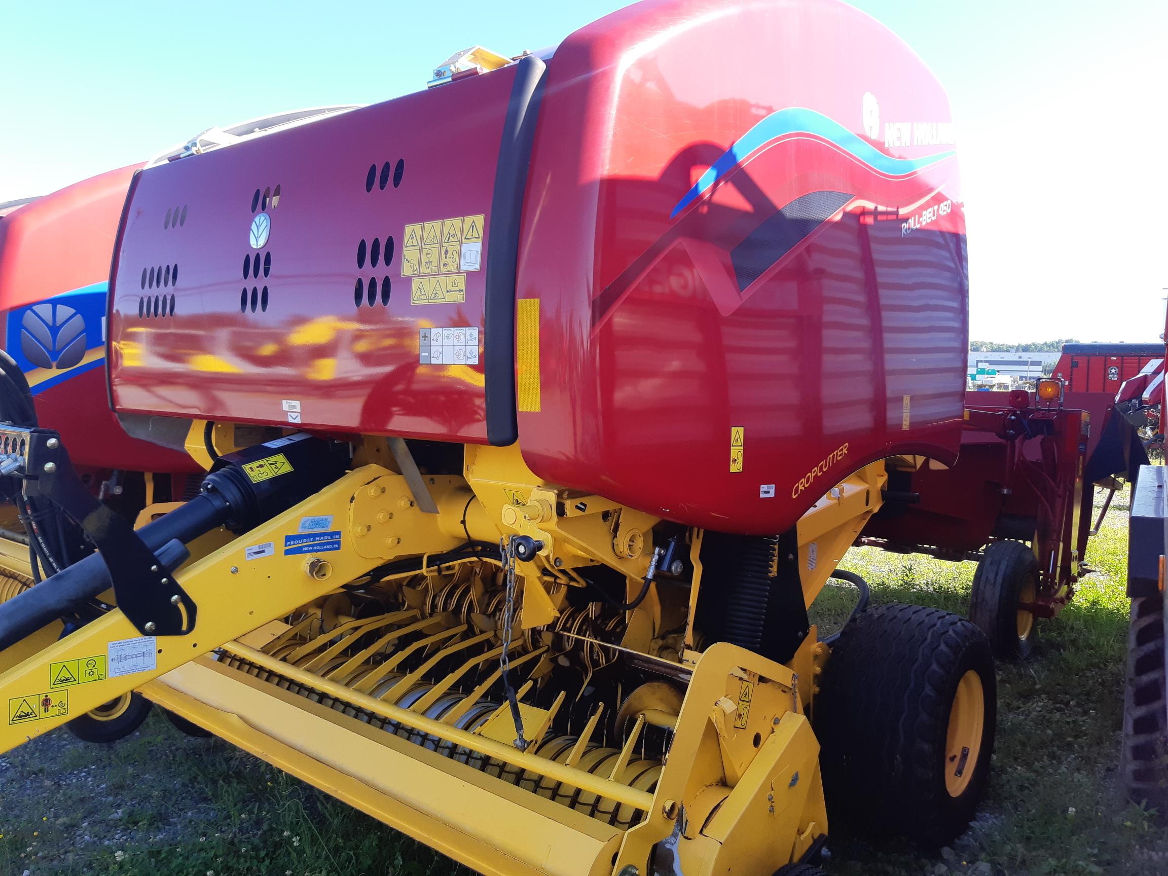 A red and yellow New Holland round baler is shown. The machine has various warning and instructional stickers on its body and is equipped with a complex assembly of metal components for baling agricultural materials.