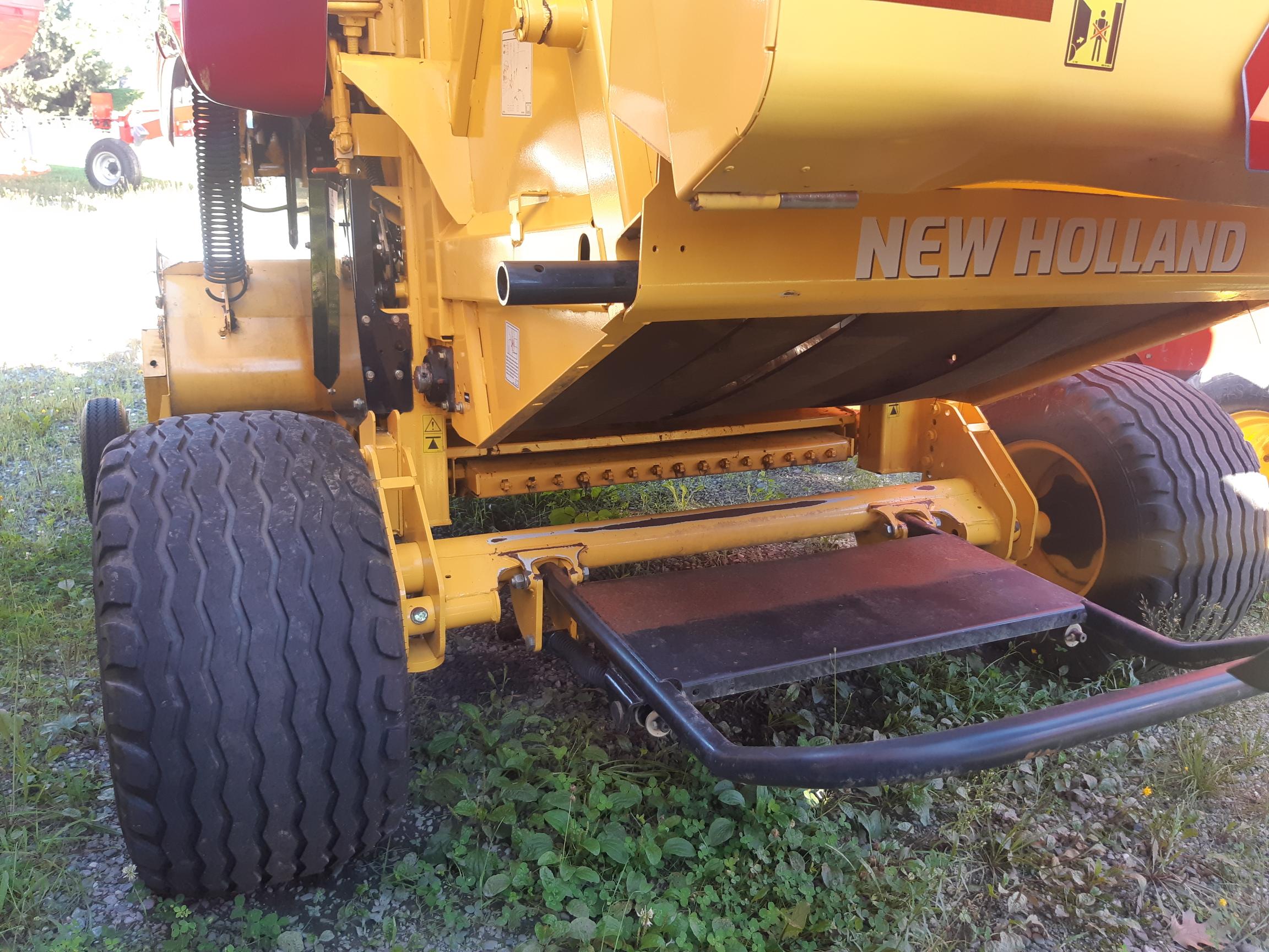 Close-up of a yellow New Holland agricultural machine, focusing on the back section. The equipment features large tires, a metal step or platform, and various mechanical components. Grass and gravel are visible on the ground around the machine.