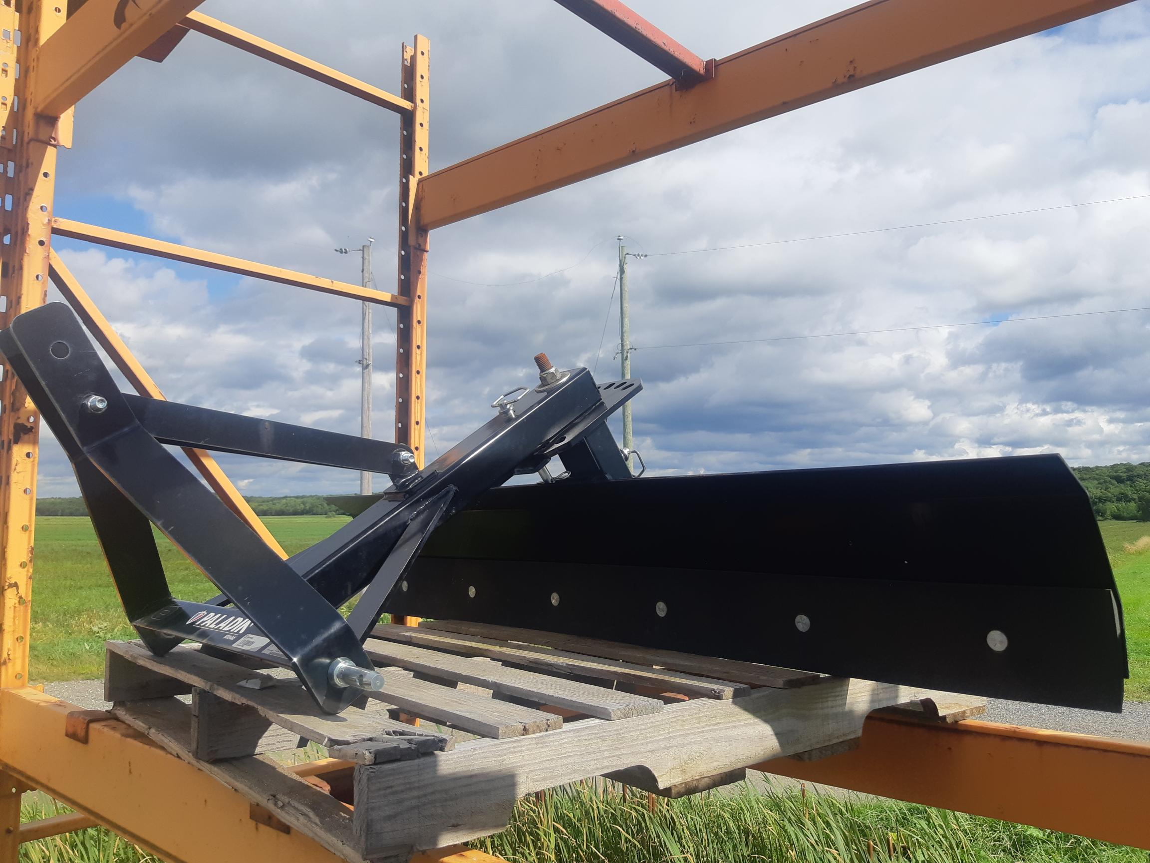 A black agricultural attachment, possibly a grader blade, mounted on a wooden pallet. The equipment is placed on an orange metal rack outdoors, with a background of grassy fields and a partly cloudy sky.