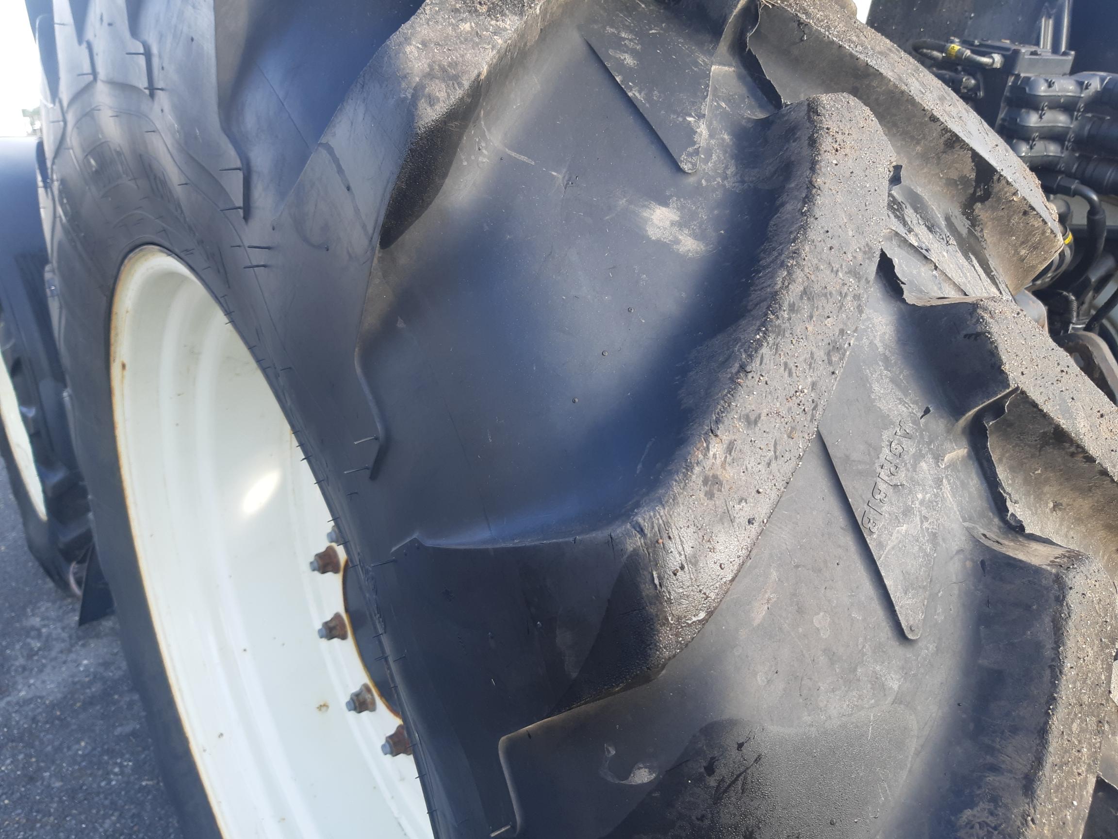 Close-up of a large tractor tire with white rims, showing significant wear and damage on the treads. The tire appears to be heavily used and possibly in need of replacement. The background is slightly blurred, but parts of the tractor's frame are visible.