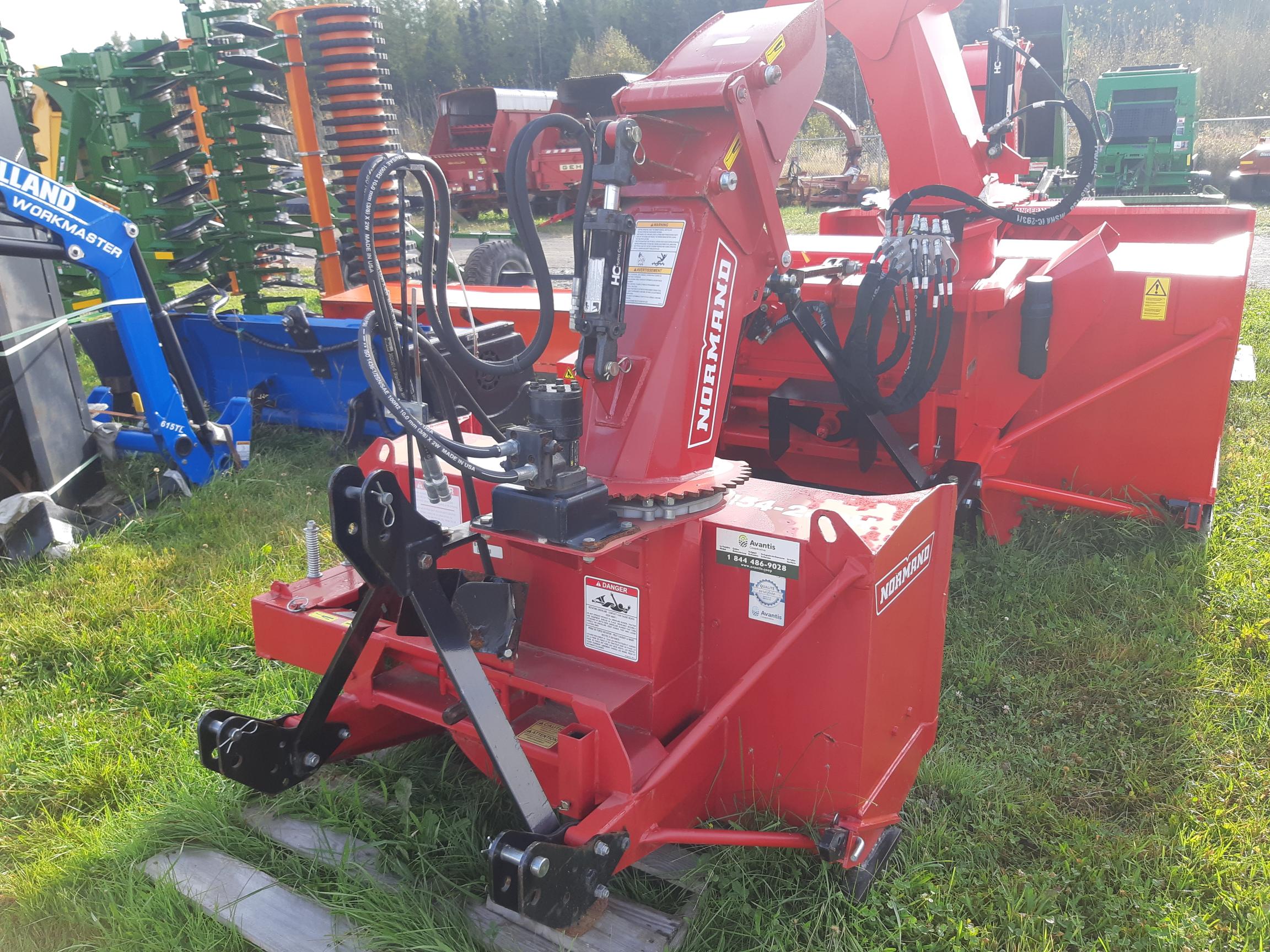 A red agricultural machine sits on a grassy field. The equipment features various levers, hydraulic hoses, and metal components. Other pieces of machinery are visible in the background, including green and blue equipment.