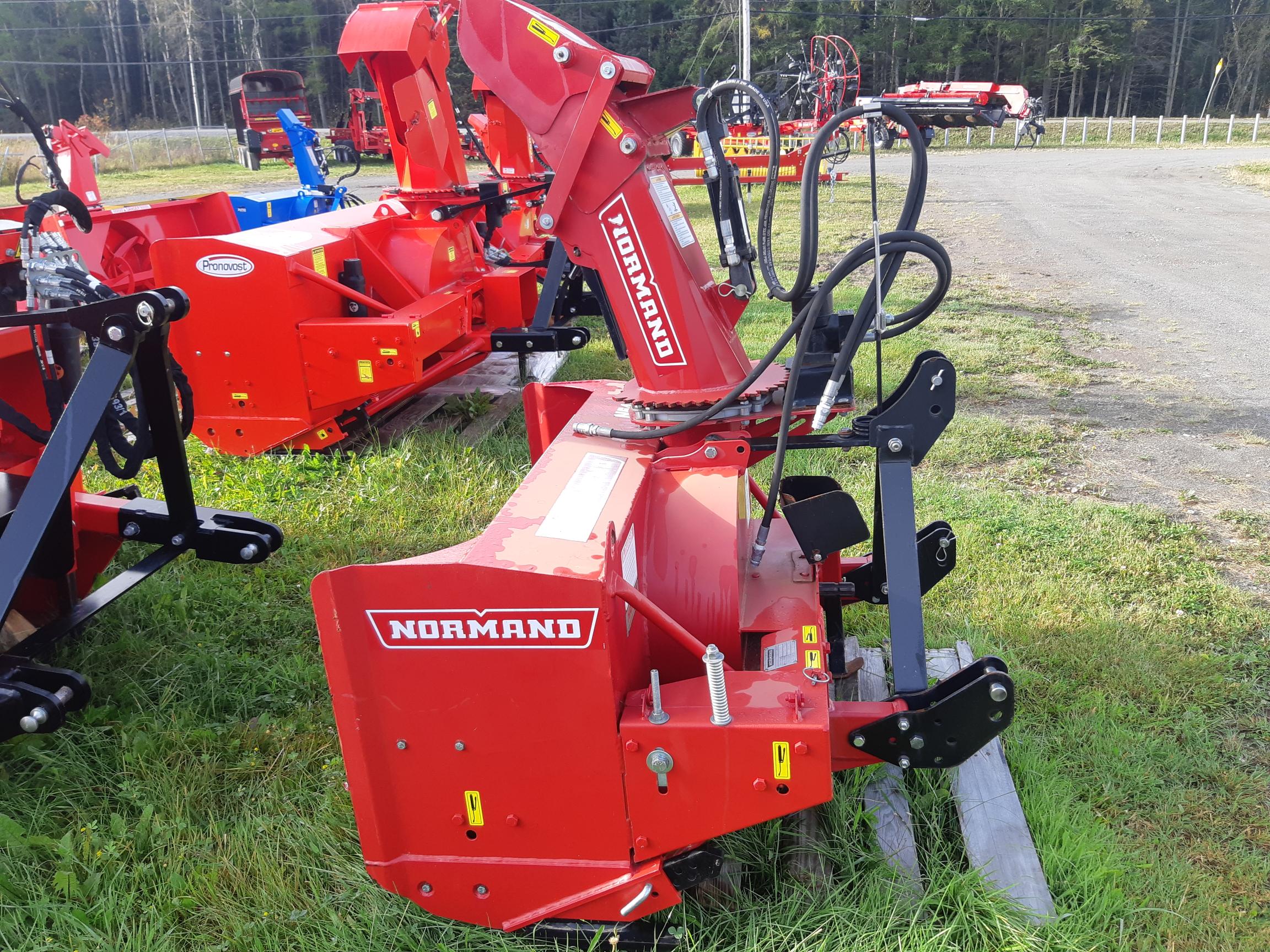 A red Normand snow blower attachment for a tractor is displayed on grass. It has a tall chute for directing the snow and various hydraulic components. Several other pieces of agricultural equipment are visible in the background, set against a wooded area and an overcast sky.