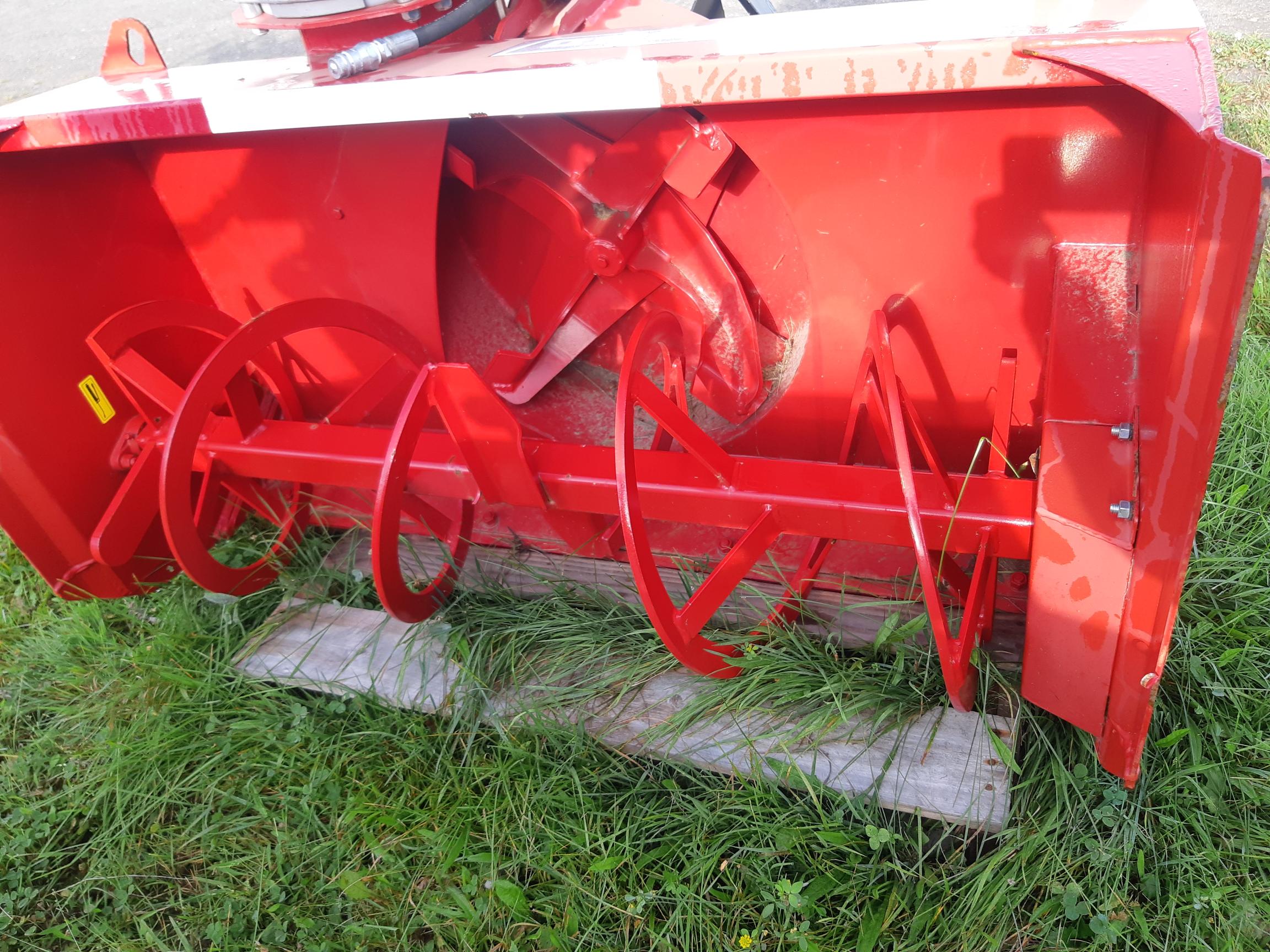 A red mechanical device with a series of rotating blades stands on a patch of grassy ground. The bright red metal shows wear marks. The grass beneath shows signs of mowing, and a wooden plank lies under the device.