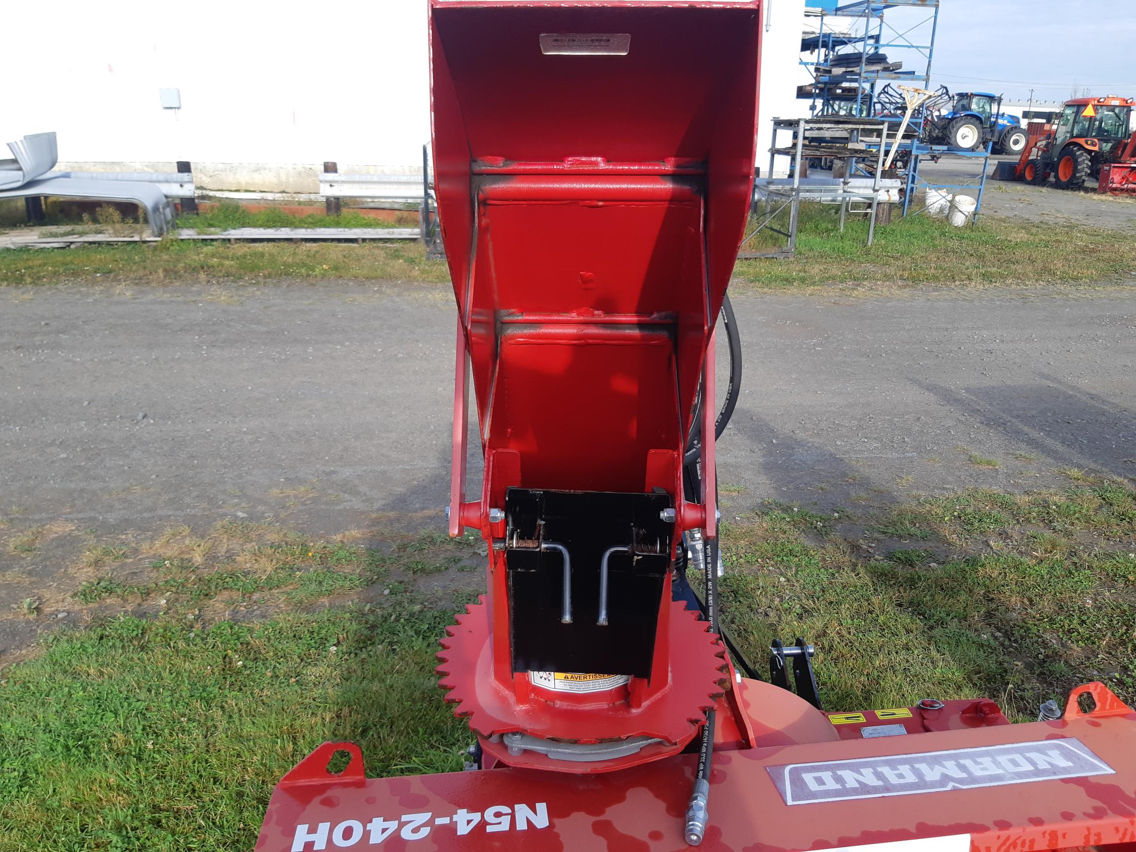 A close-up view of a red Kubrand NFM-240 flail mower attachment on a dirt and grass field. The image shows the feed opening and part of the machine's mechanism. Farm equipment and a building are visible in the background under a clear sky.