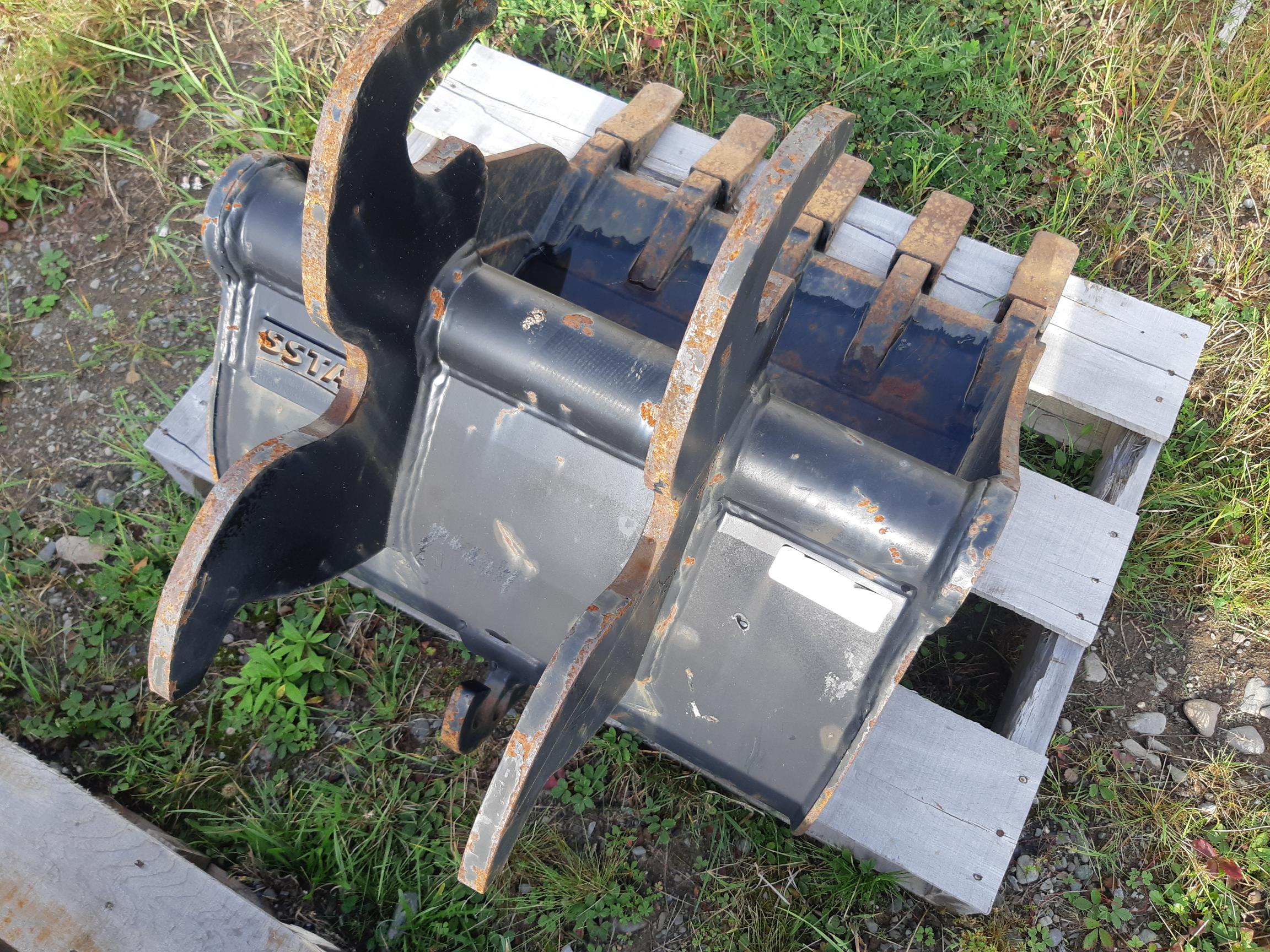 A heavy-duty excavator bucket attachment rests on a wooden pallet outdoors. The dark metal bucket shows signs of wear with rust patches and dirt, indicating frequent use. Surrounding the pallet is grass and small plants among a rough, uneven ground.