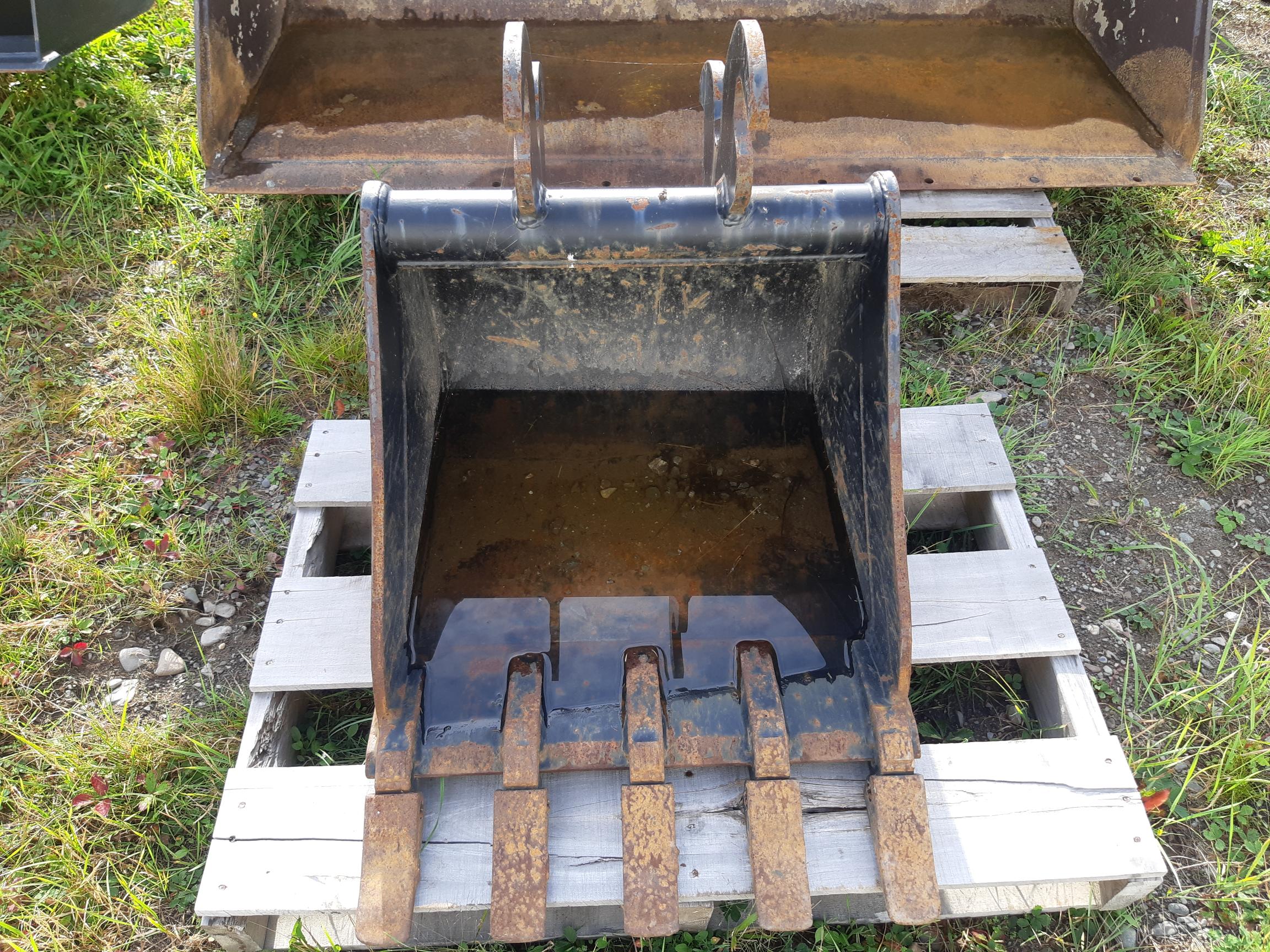 A rusty excavator bucket attachment rests on a wooden pallet outdoors. The bucket has five metal teeth and visible wear and tear. Surrounding the pallet is grass and gravel. Another similar attachment is partially visible in the background.