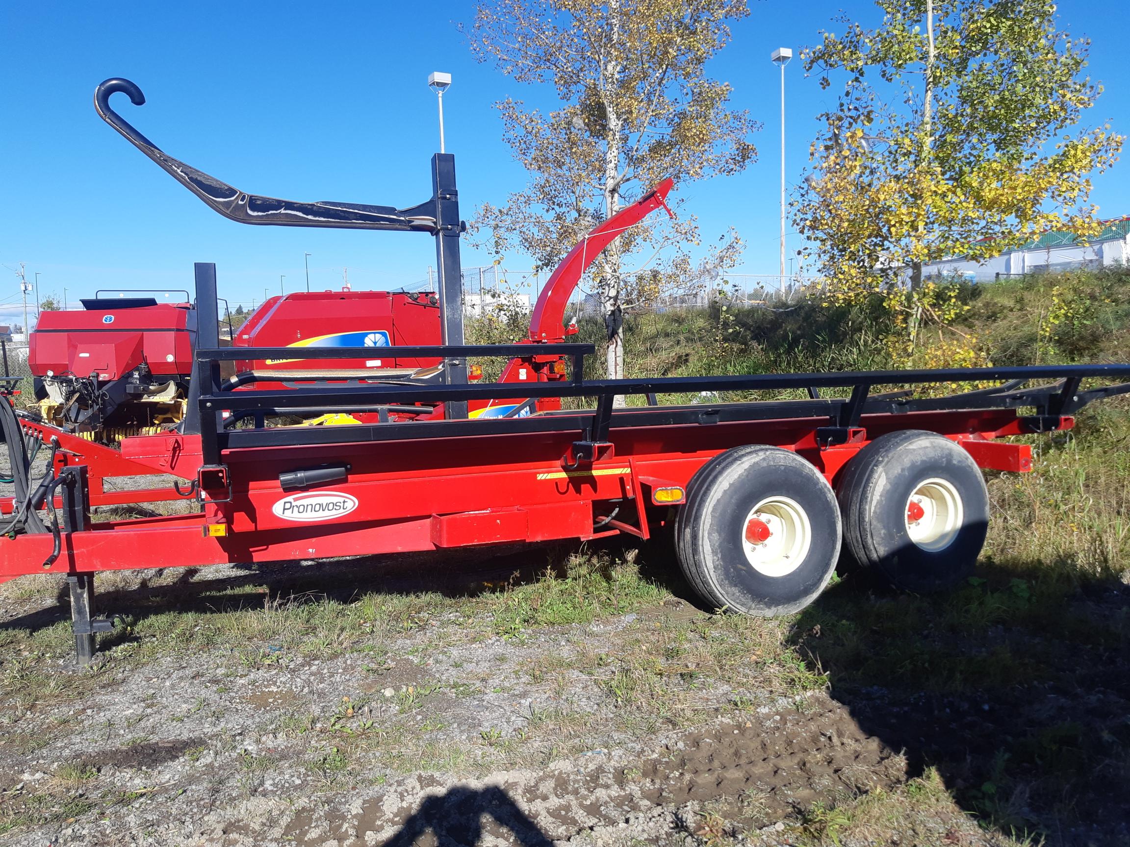 A red agricultural trailer with two large wheels and a mechanical arm attachment, designed for transporting and handling hay bales, is parked on a grassy area. Bright blue sky and trees with some yellow leaves are visible in the background.