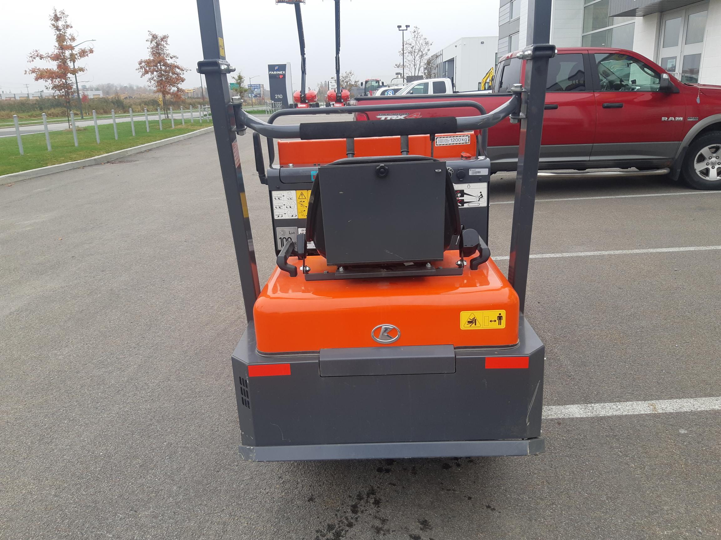 A compact, orange construction vehicle with two black roll bars and control panel is parked in a lot. To the right, there is a red pickup truck and a building in the background. The pavement is bordered by a grassy area and a row of leafless trees.