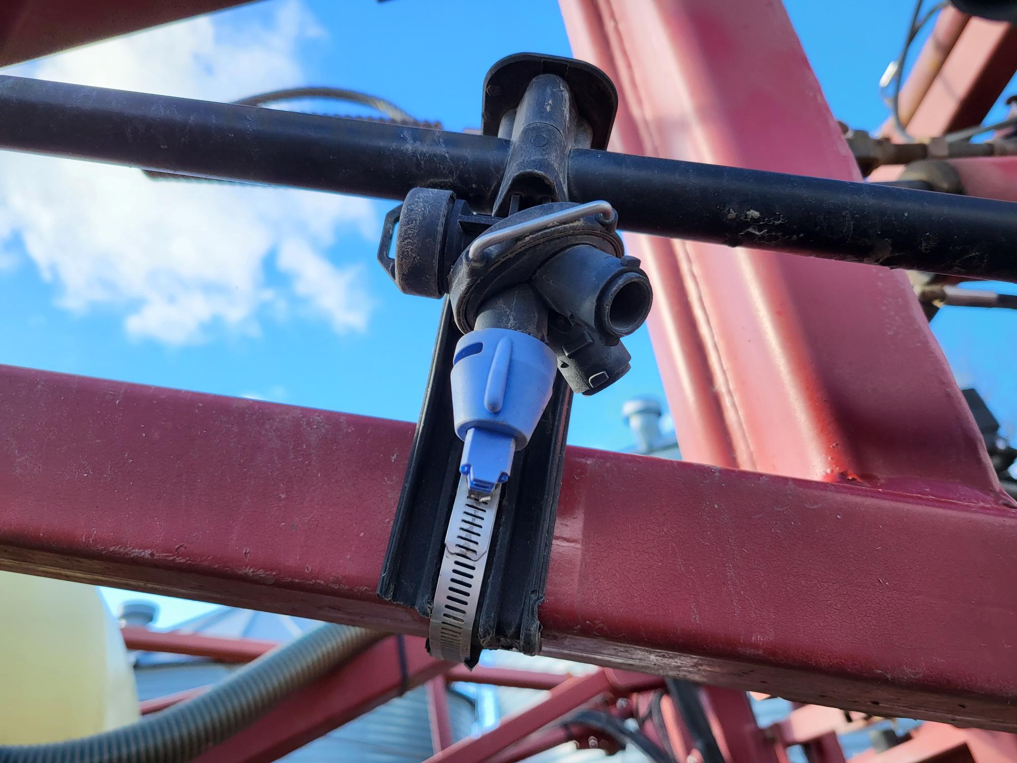 Close-up of a plastic nozzle attached to a black pipe, mounted on a red metal frame. The nozzle’s blue cap and surrounding fittings are clearly visible against a bright blue sky with a few clouds. Background shows parts of machinery and a distant building.