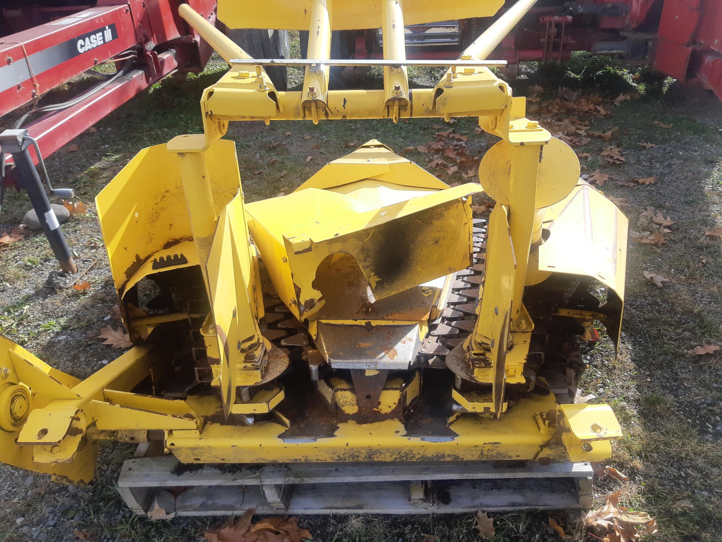 Yellow agricultural machinery attachment, possibly a crop harvester, with visible gears and blades, standing on wooden pallets outdoors. Red machinery and fallen leaves are in the background. The machine appears weathered and rusty in some areas.