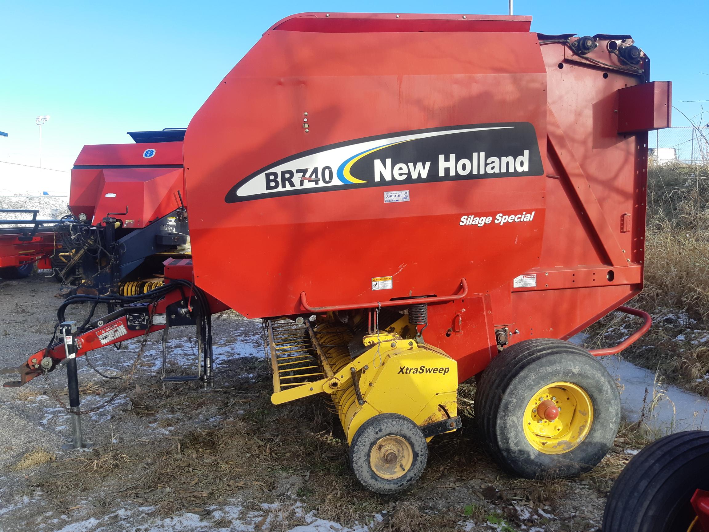 The image shows a New Holland BR740 Silage Special round baler. It is predominantly red with black, yellow, and white components. The equipment is situated outdoors on ground with patches of snow and hay in the surroundings.