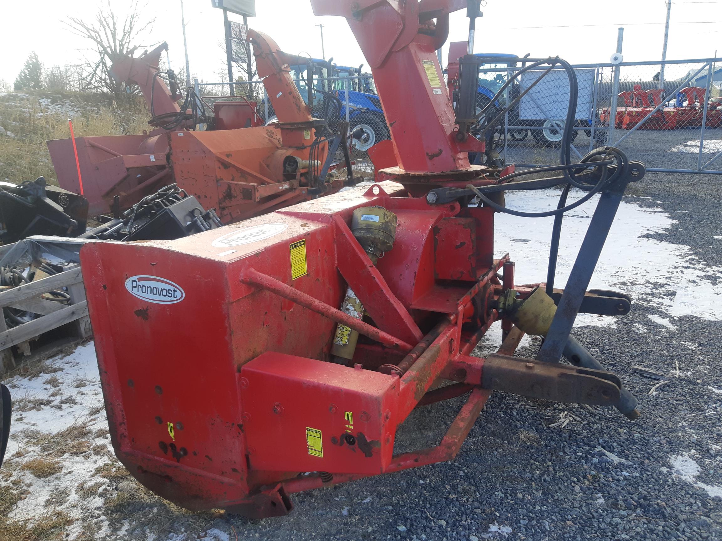 A red Pronovost snow blower attachment is positioned outdoors on a gravel lot with patches of snow. The equipment is rusted and shows signs of wear. Other similar machinery and blue fencing can be seen in the background along with some bare trees.