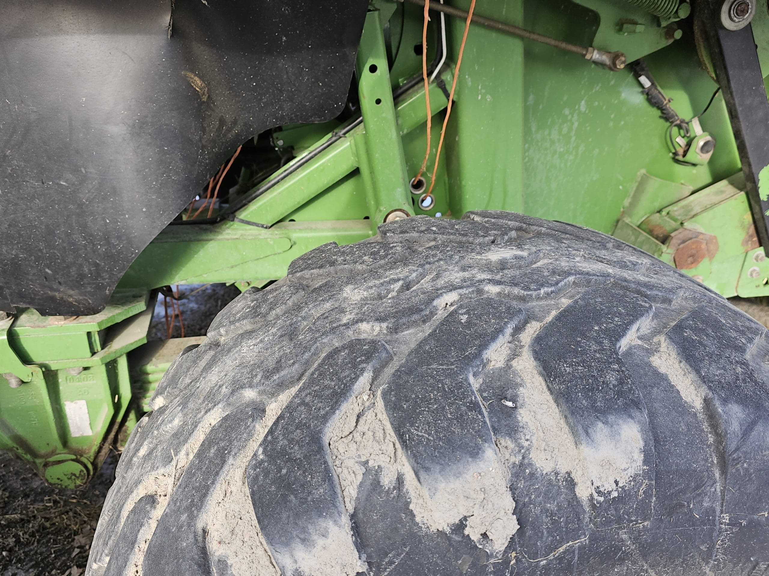 Close-up of a large tractor tire partially covered in mud. The tire is attached to green metal machinery with some orange cables visible. The background includes part of the tractor's chassis and components.