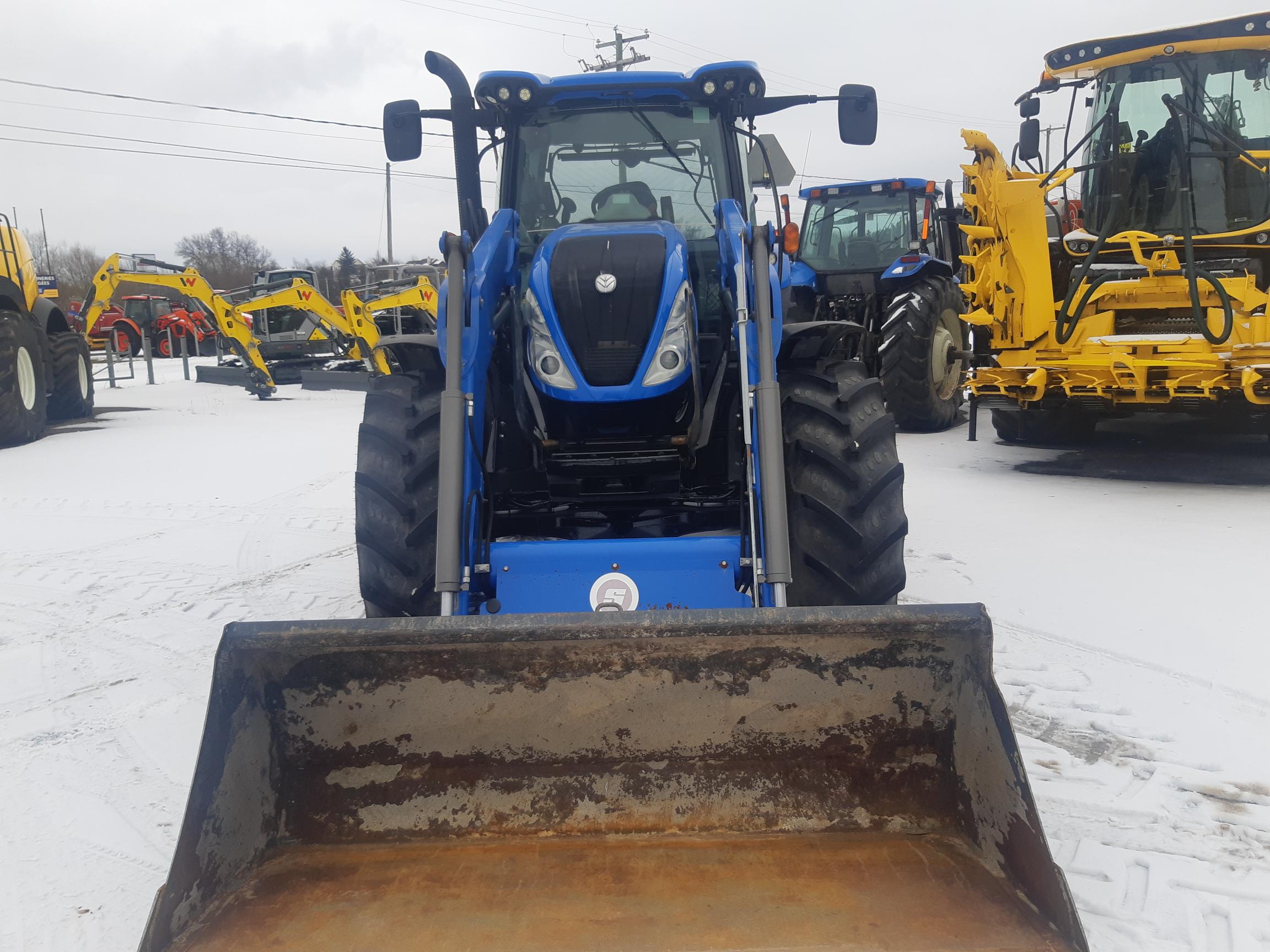 A blue tractor with a front loader attachment is parked in a snowy lot. Other tractors and yellow construction equipment are visible in the background. The sky is overcast, and the ground is covered with snow.