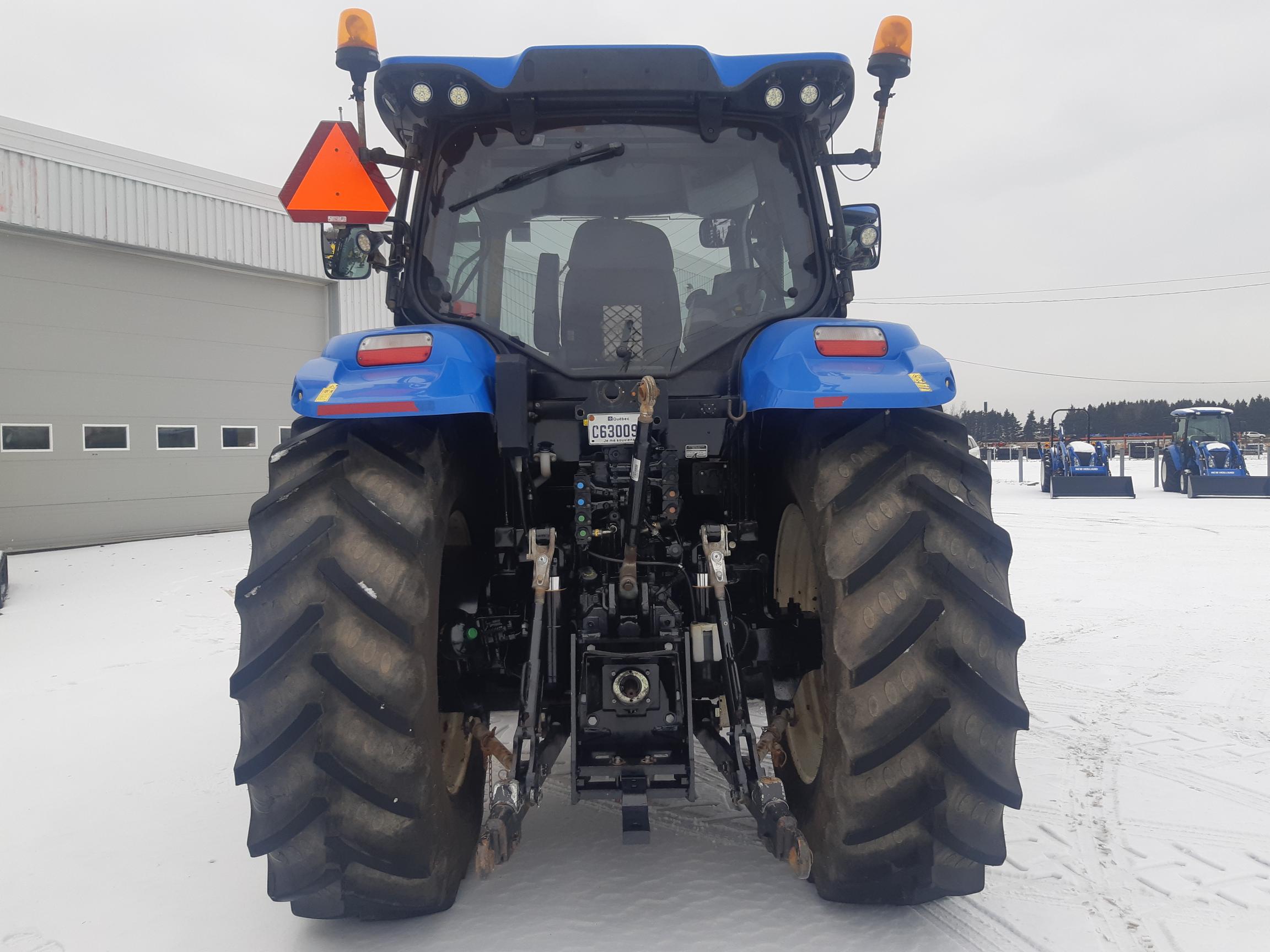 A large blue tractor is seen from the rear, showcasing its substantial tires and various mechanical components. It features bright orange hazard lights on top and a red reflective triangle. Snow covers the ground, and several other tractors are visible in the background.