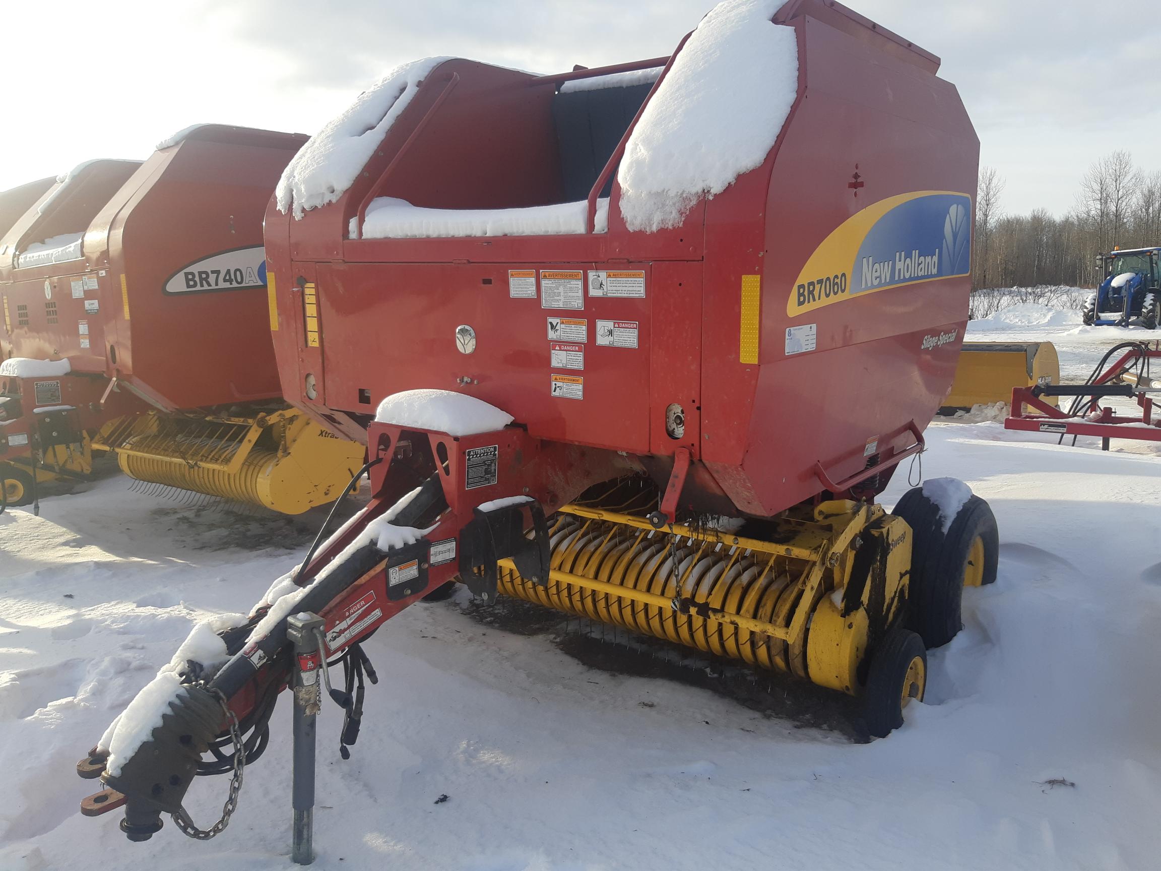 A red New Holland BR7060 round baler is parked in a snowy field. The machine, used for baling hay, is partially covered in snow. Another red baler is visible in the background. Trees and a tractor are in the distant background under a cloudy sky.