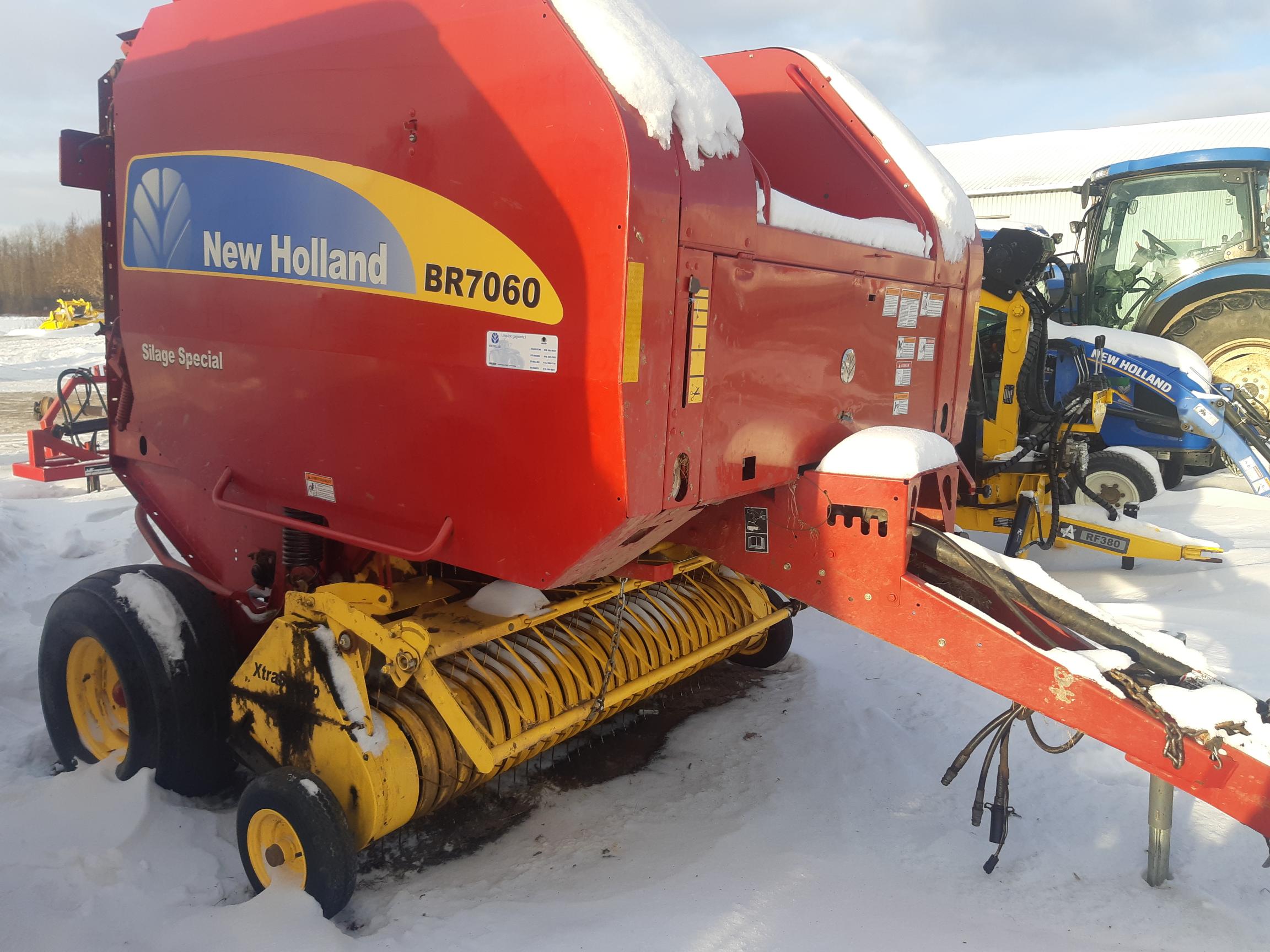 A red New Holland BR7060 baler is parked in a snowy area with a yellow front component. The baler has the phrase 